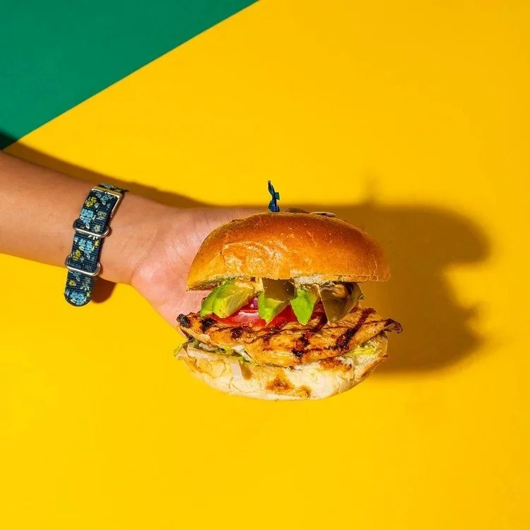 Hand holding an Empanada Mama grilled chicken sandwich layered with vegetables against a bold yellow background.