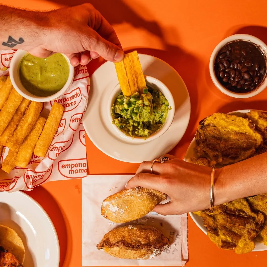 A hand reaches for Empanada Mama’s golden empanadas beside guacamole and green sauce.