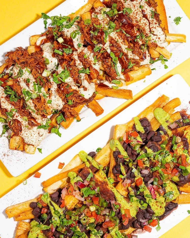 Loaded fries from Empanada Mama topped with pulled meat, beans, fresh salsa, and creamy sauces served on a white platter.