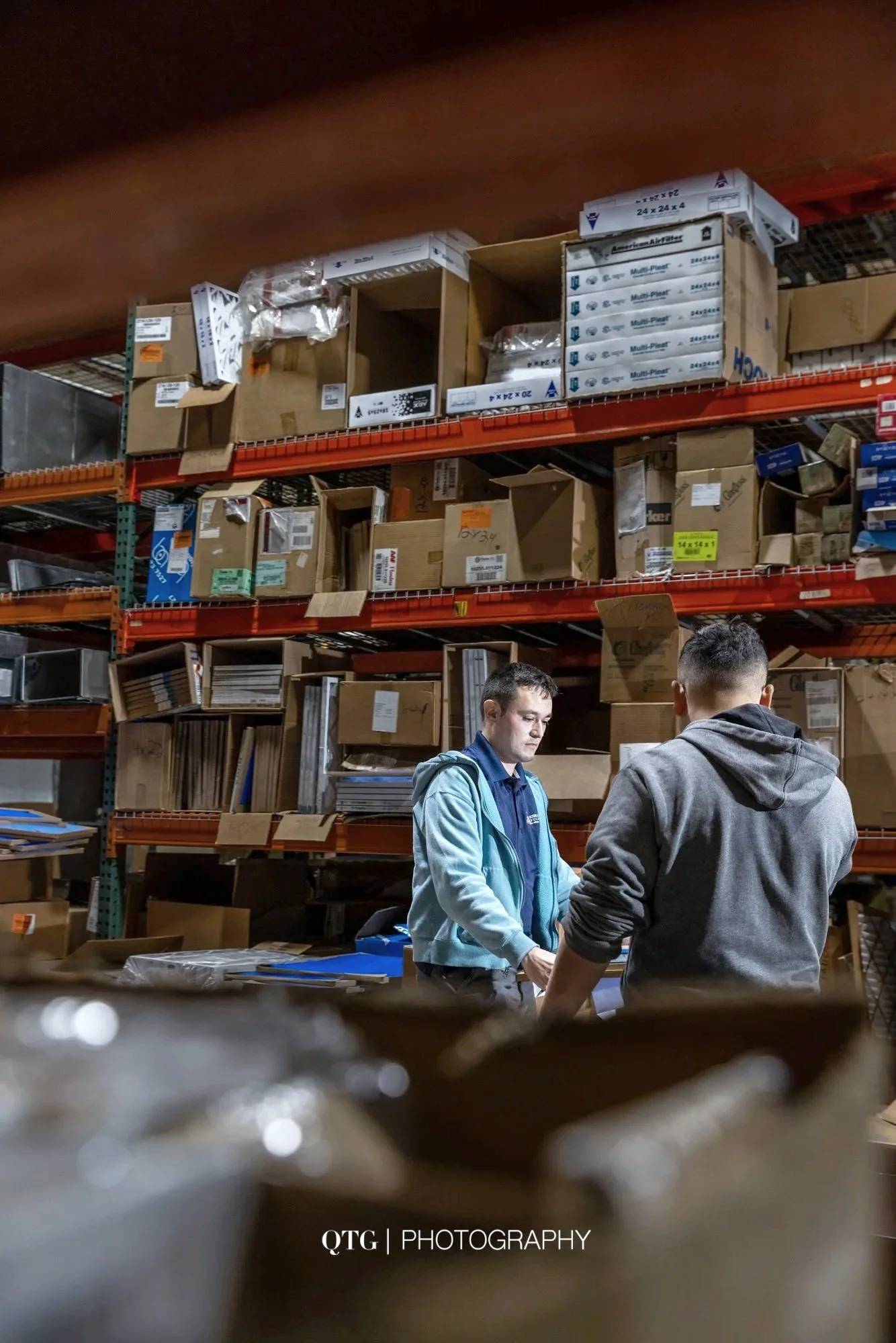 Two people engaging in a conversation in a warehouse or storage room filled with boxes and shelves of packages.