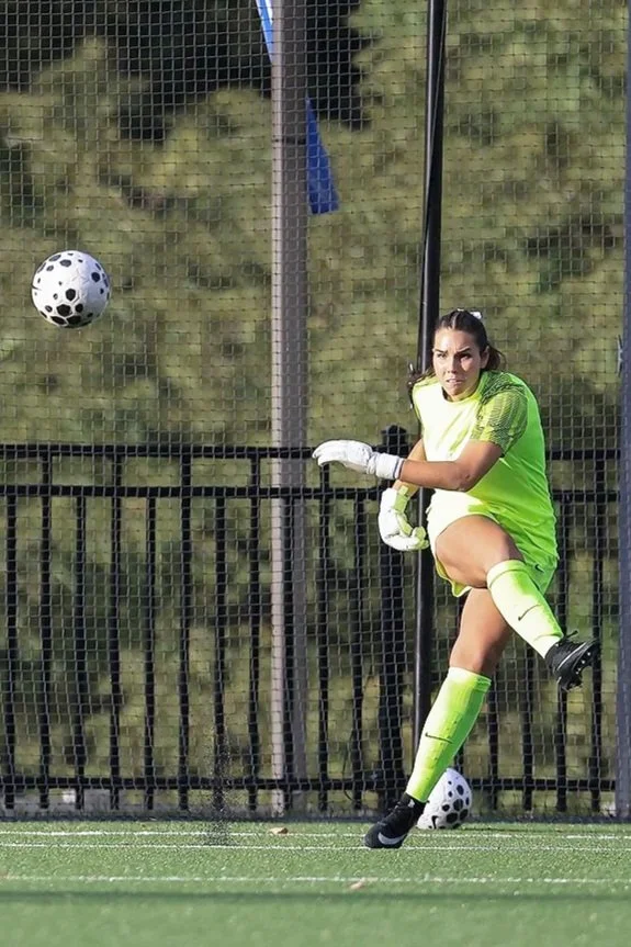 Female soccer goalkeeper in a neon yellow uniform preparing to block a soccer ball during a game on a green field, with a black fence and trees in the background.