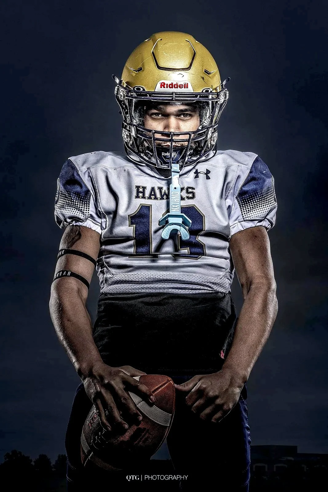 Young male football player wearing helmet and uniform, holding a football, standing against a dark background.