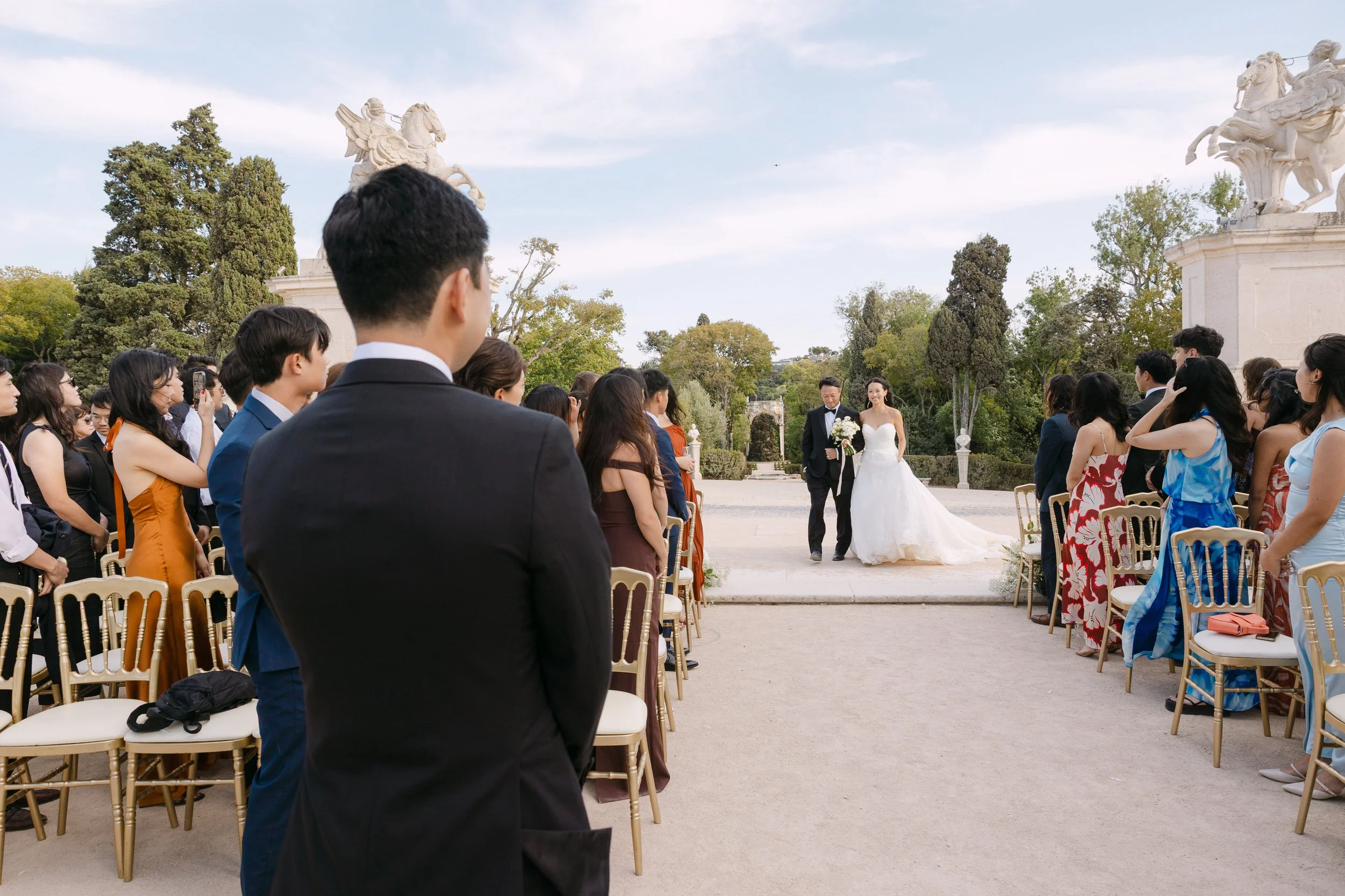 Bride arriving at the outdoor ceremony as the groom watches from the aisle, surrounded by wedding guests in the gardens of Palácio Nacional de Queluz.