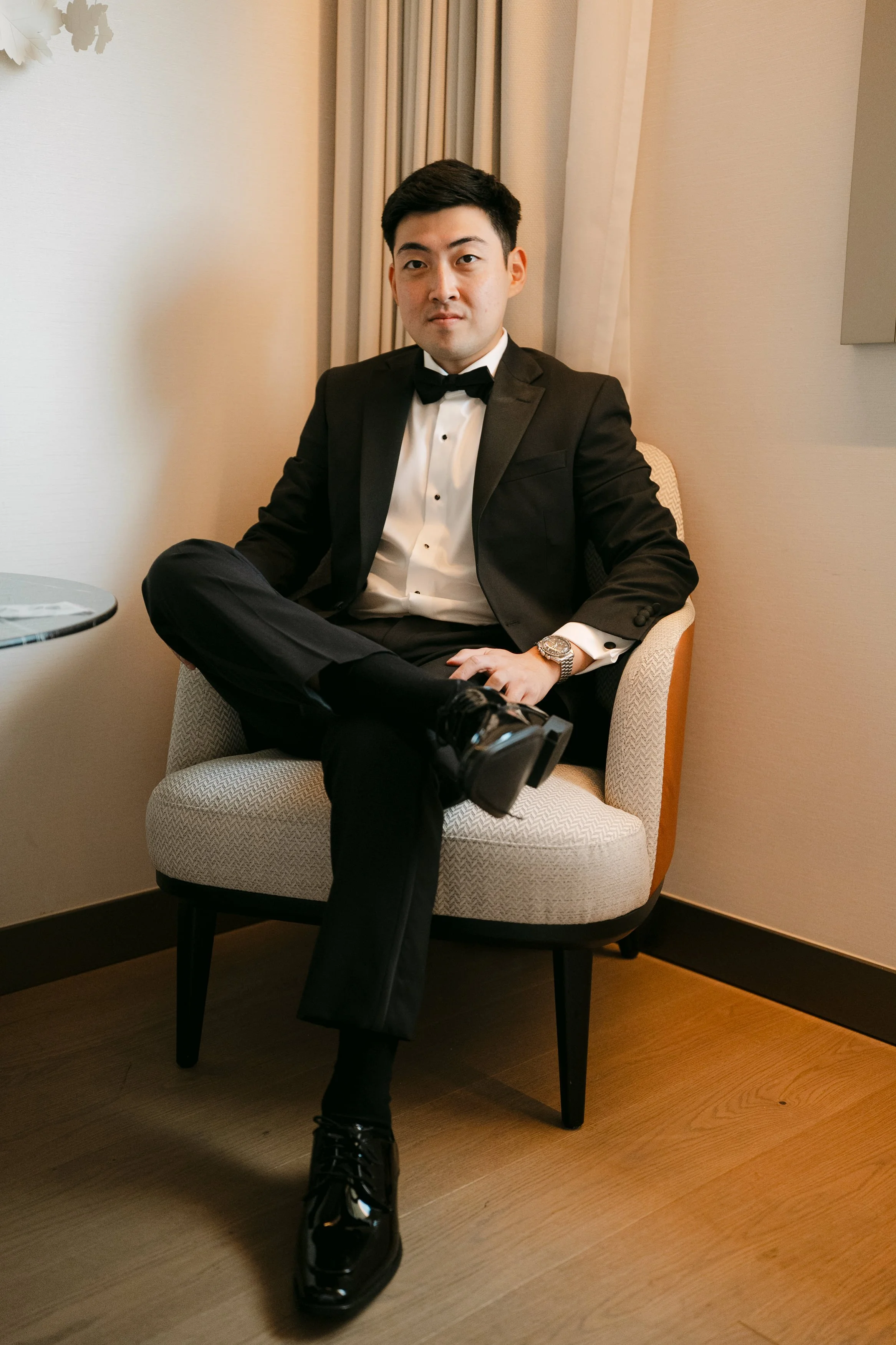 Groom wearing a classic black tuxedo and bow tie seated in an armchair before his wedding at Palácio Nacional de Queluz.