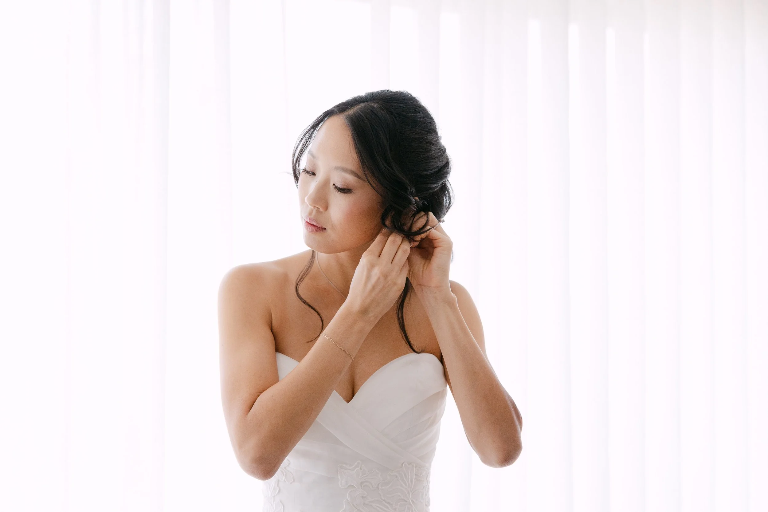 Bride putting on her earrings beside a softly lit window while getting ready for her wedding at Palácio Nacional de Queluz.