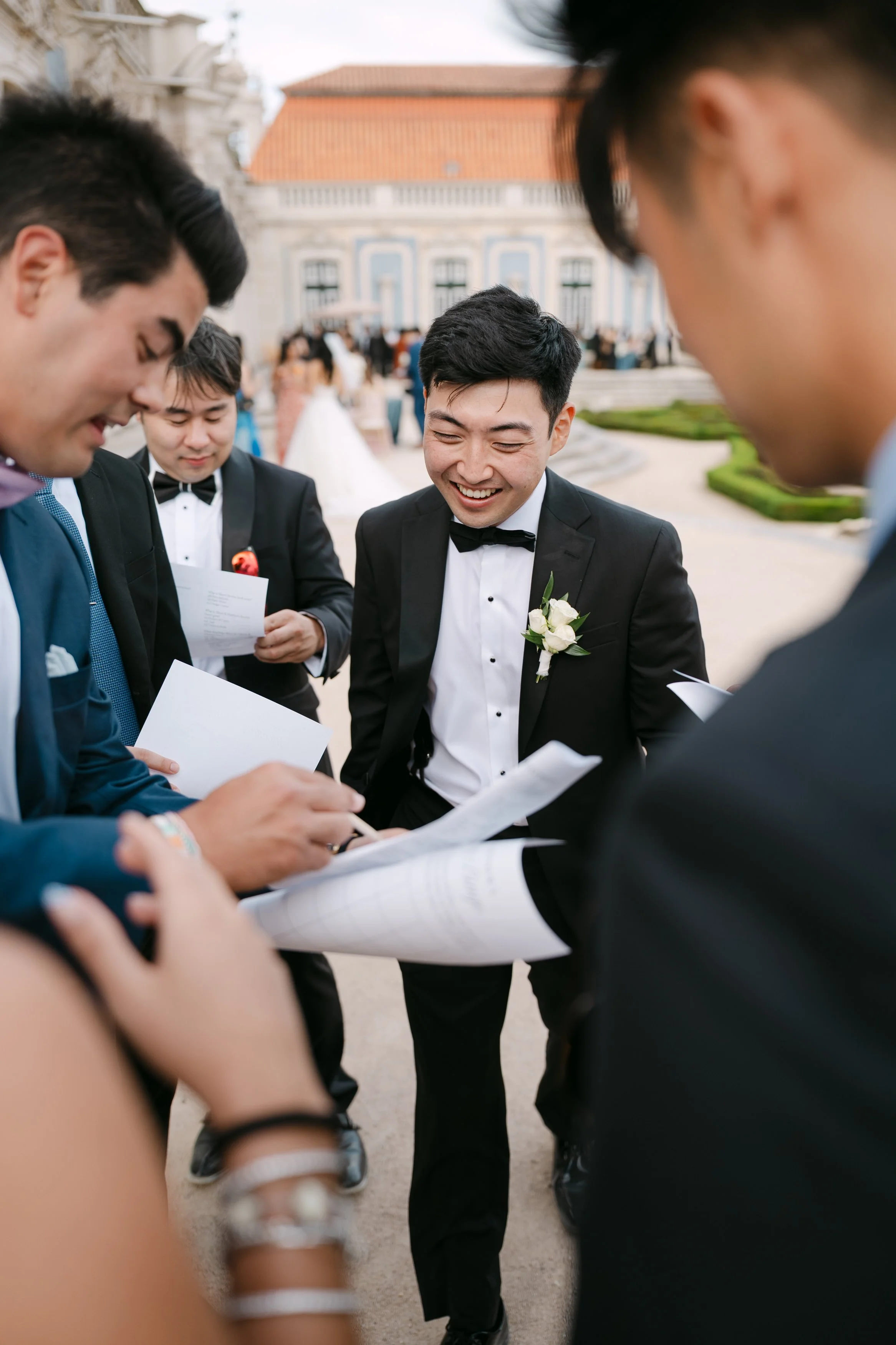 Groom laughing with friends as they read cards together during cocktail hour in the gardens of Palácio Nacional de Queluz.