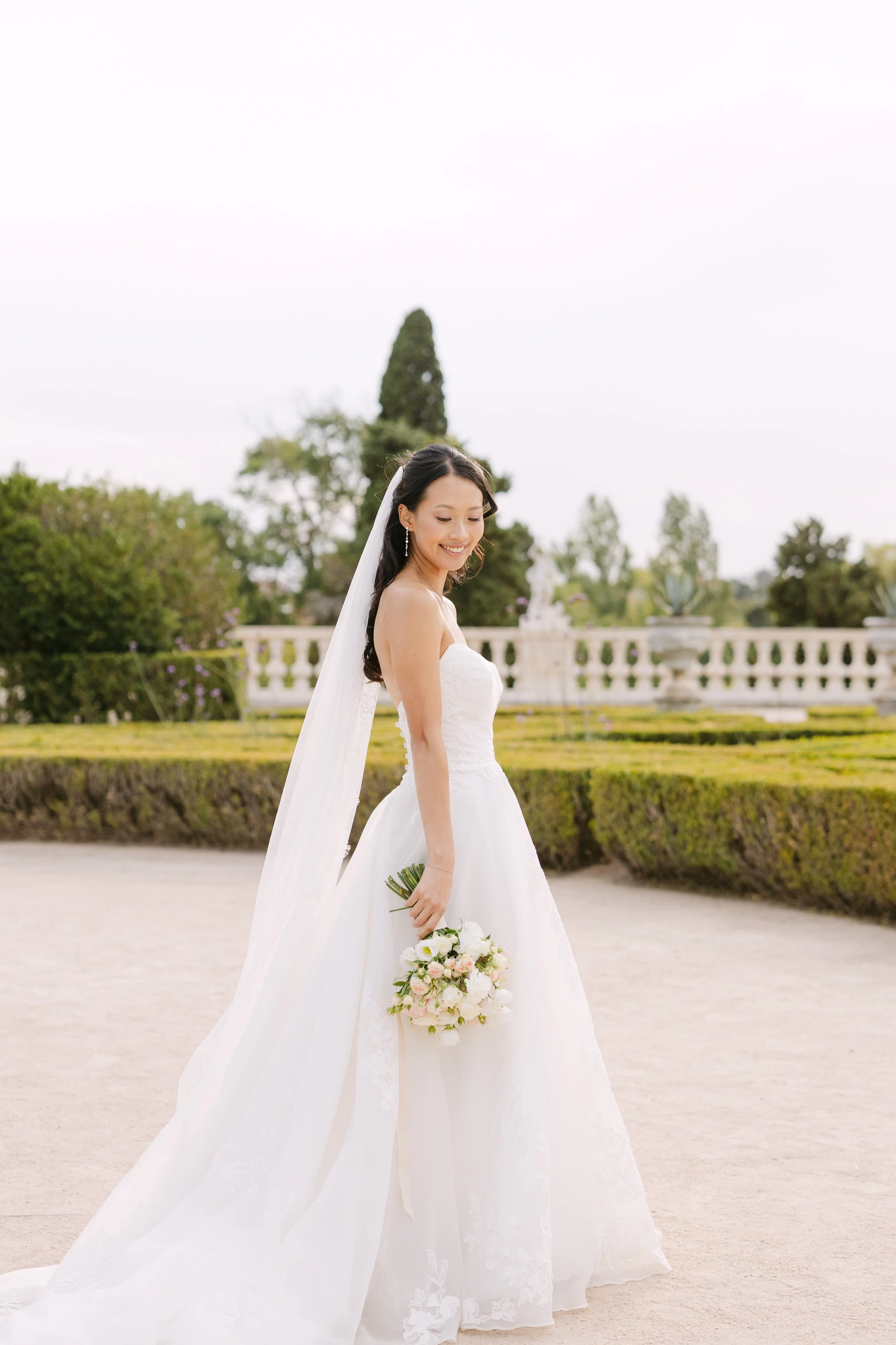 Bride walking through the formal gardens of Palácio Nacional de Queluz in her flowing white wedding gown and long veil, carrying a soft pastel bridal bouquet.