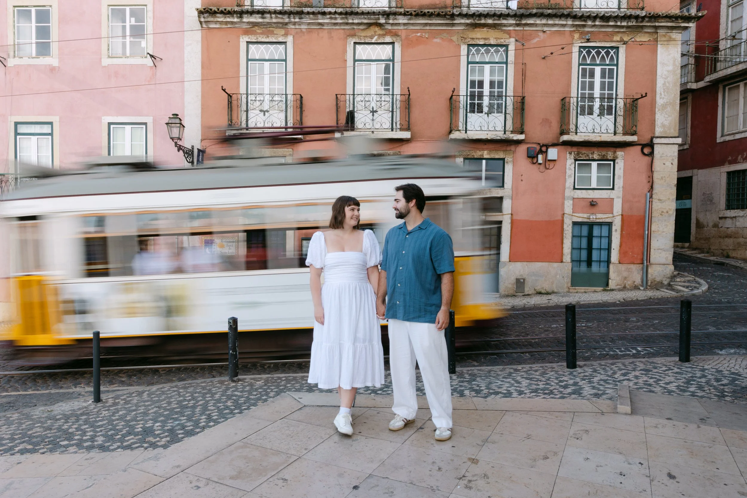Newly engaged couple photographed in Alfama with a traditional Lisbon tram passing behind them after their surprise proposal.