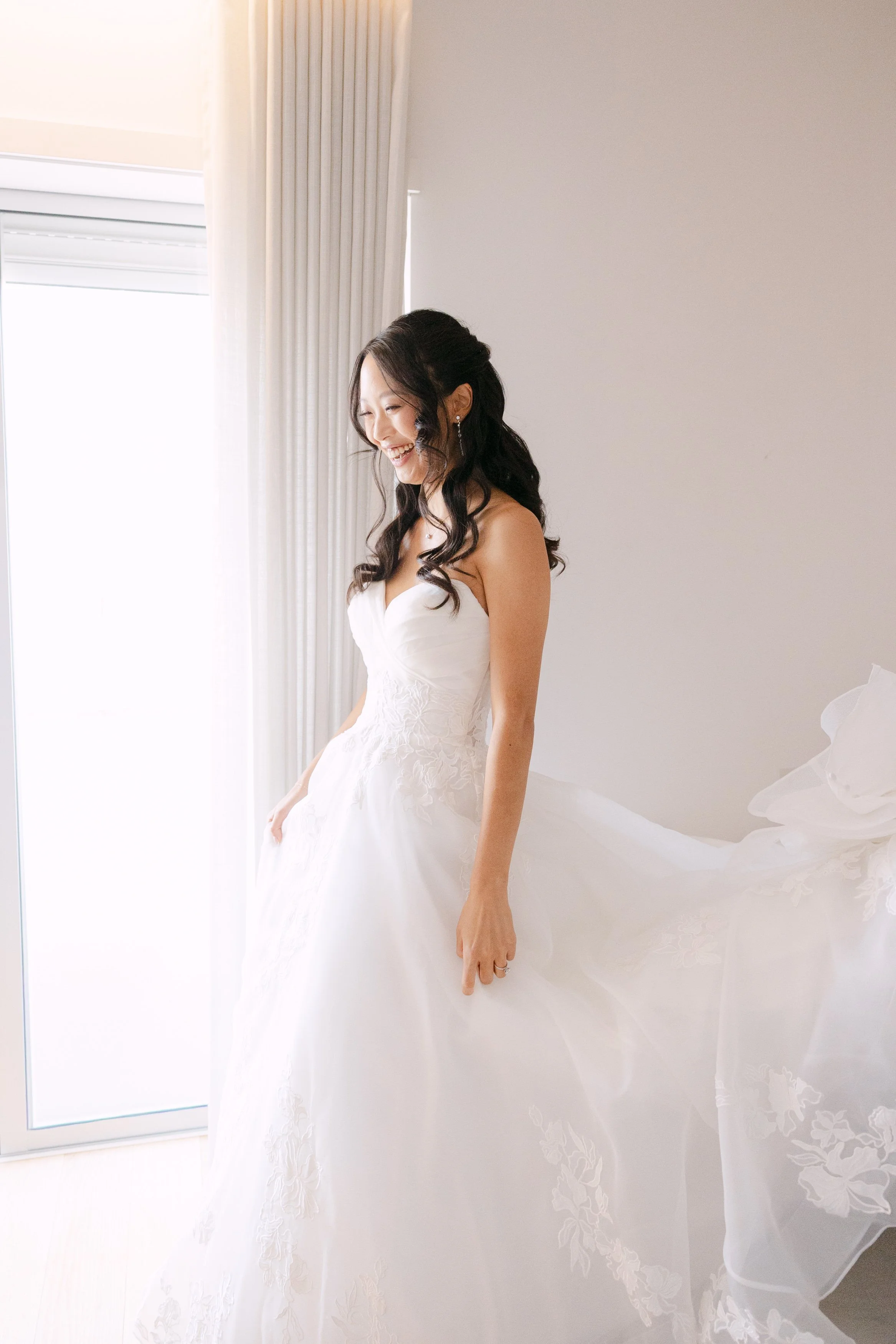 Bride standing beside a bright window in her white embroidered wedding gown during morning preparations for her destination wedding in Portugal.