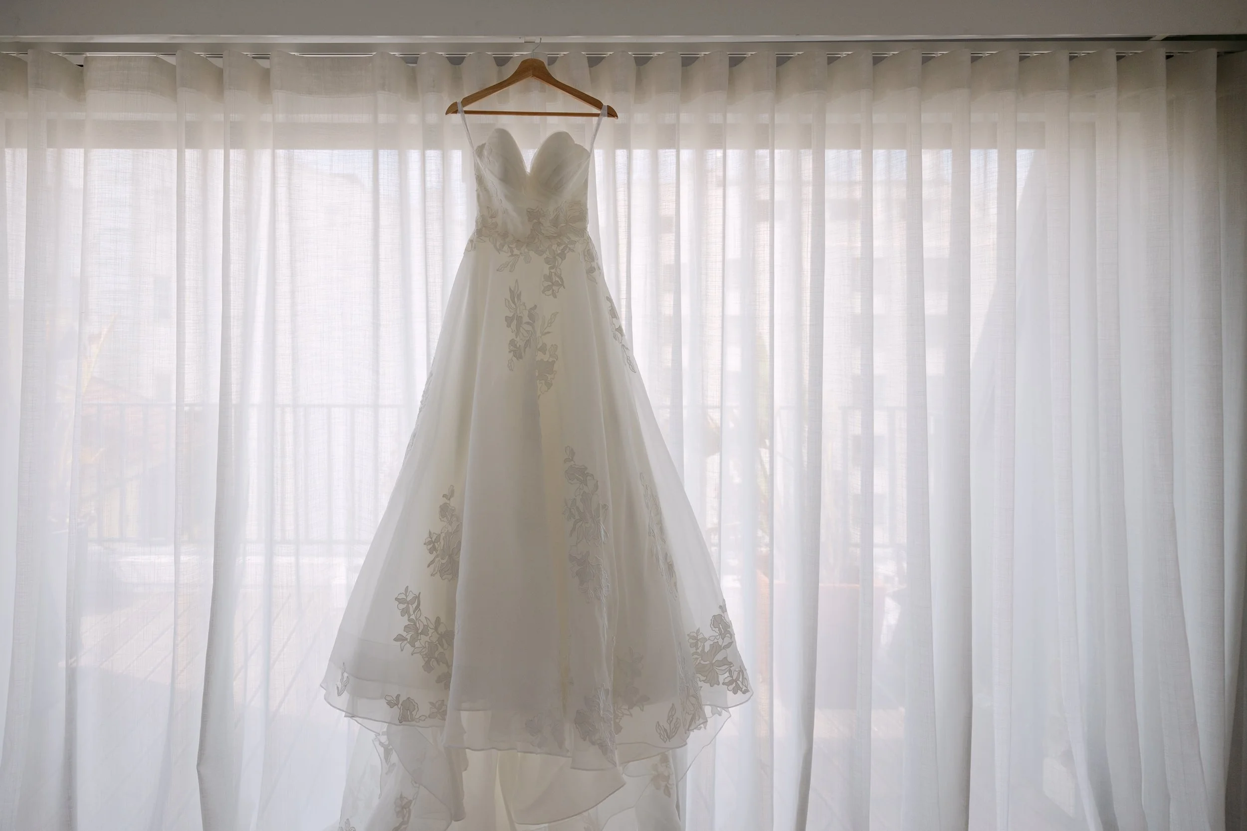 White wedding dress with delicate floral embroidery hanging in front of sheer curtains, softly backlit by natural morning light during wedding preparations in Lisbon, Portugal.