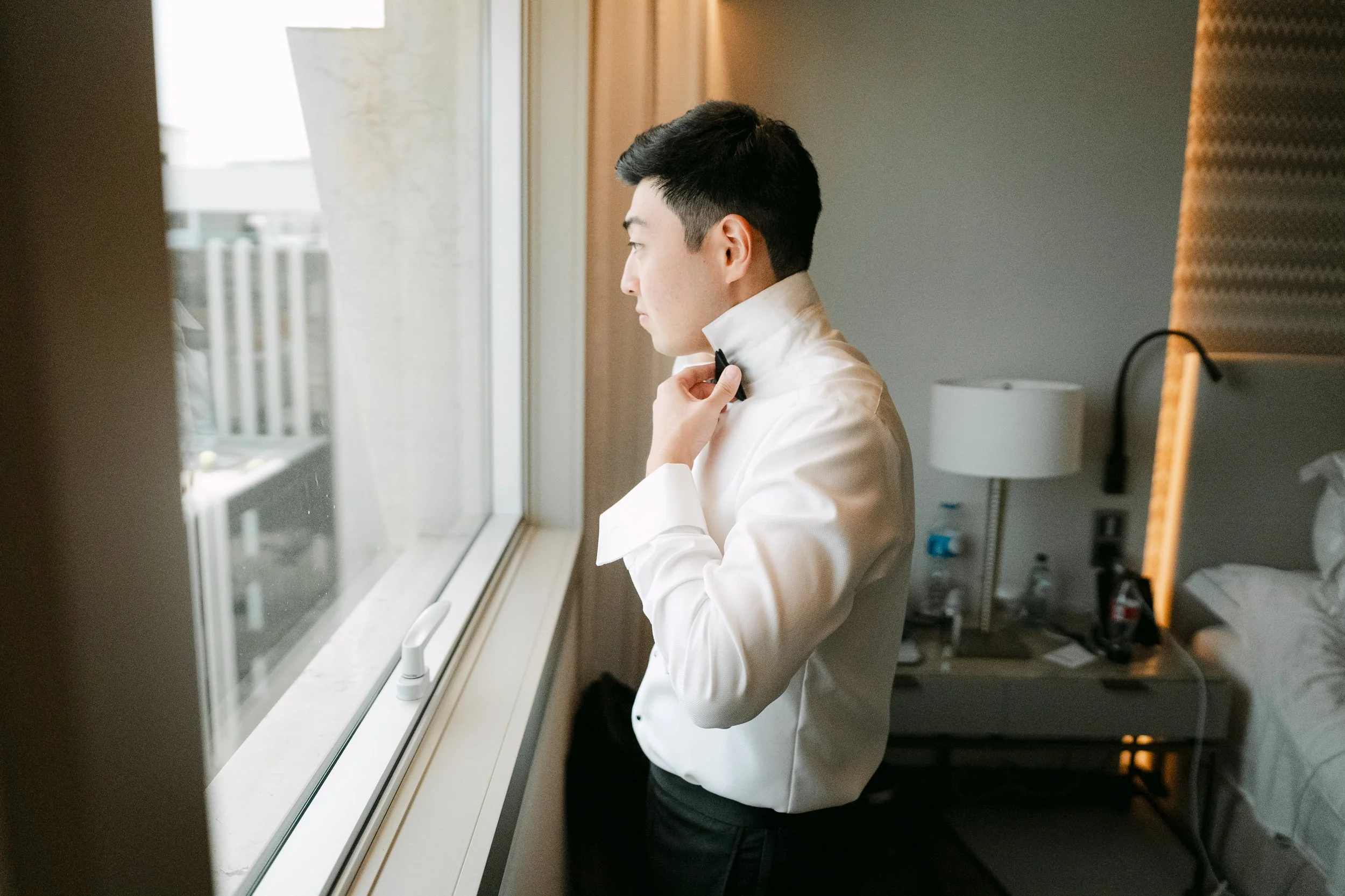 Groom adjusting his bow tie beside a hotel window while getting ready for his wedding day in Lisbon, Portugal.