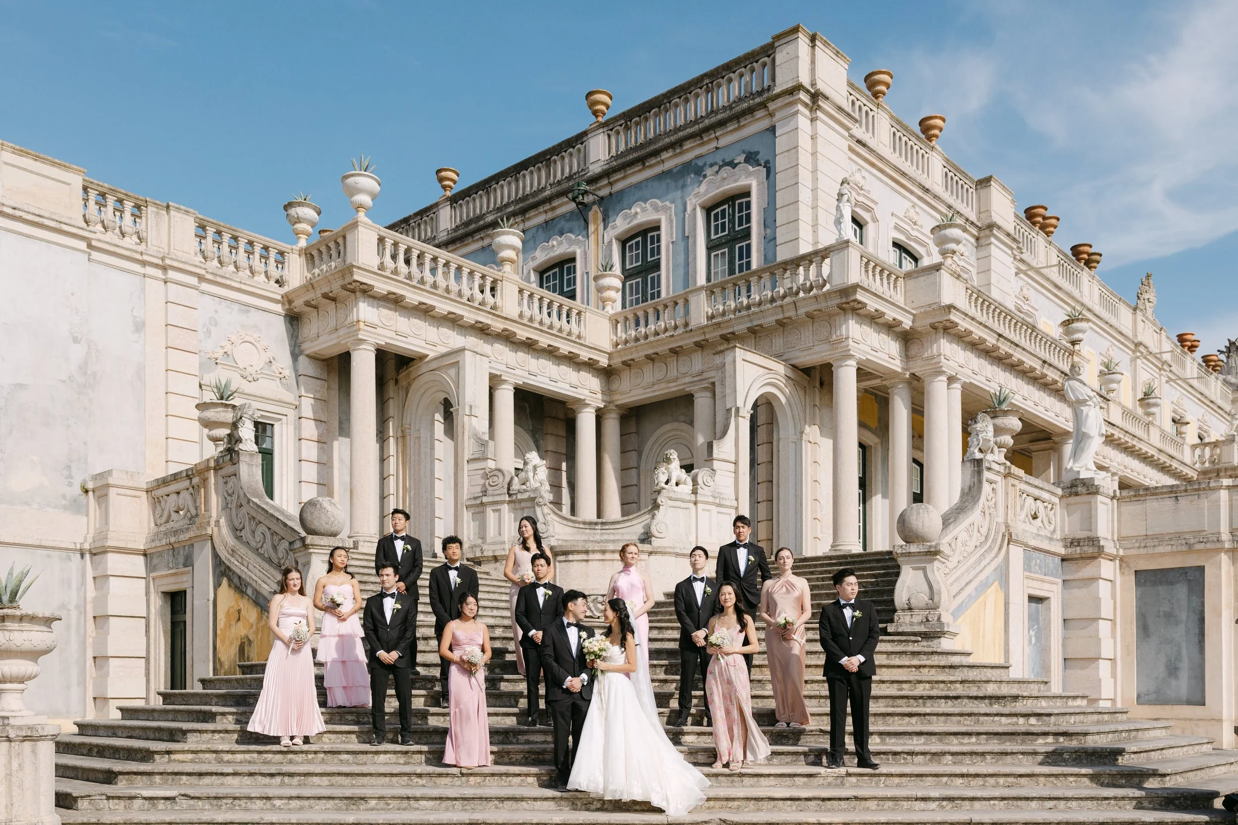 Bride and groom surrounded by their full wedding party in pink dresses and black tuxedos on the monumental staircase of Palácio Nacional de Queluz beneath a bright blue sky.