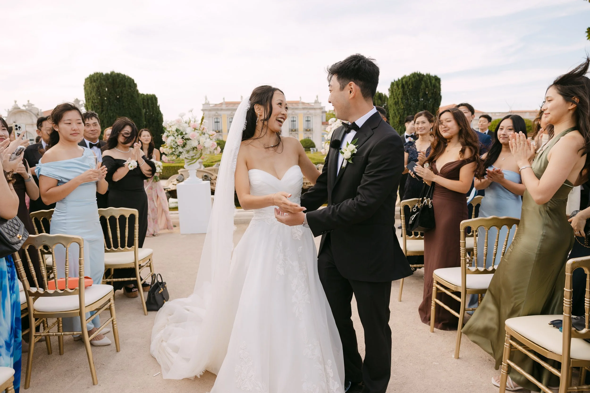 Newly married couple laughing and holding hands as they walk back down the aisle while their guests applaud after the ceremony.