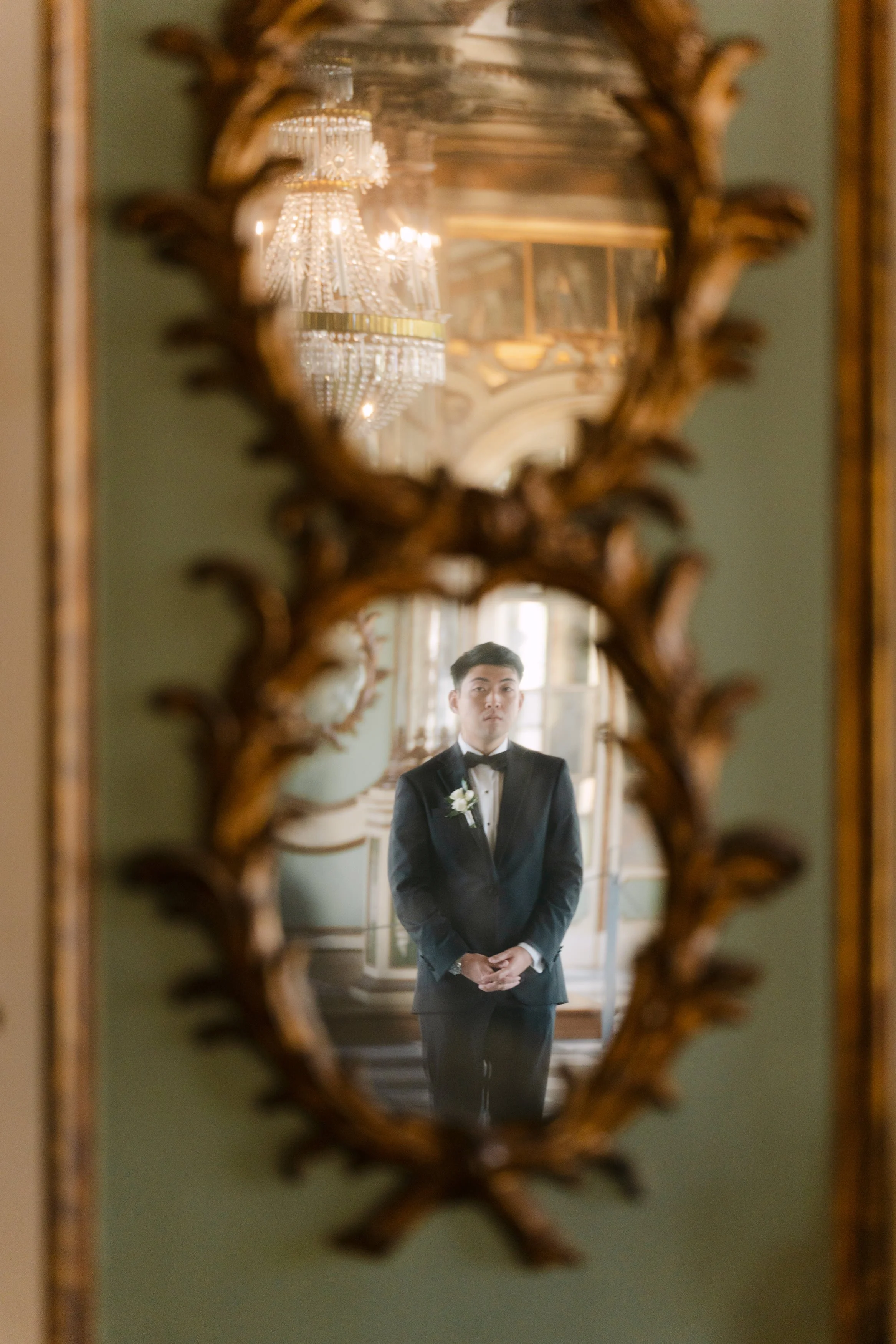 Groom in a black tuxedo reflected in an ornate antique mirror inside the historic rooms of Palácio Nacional de Queluz.
