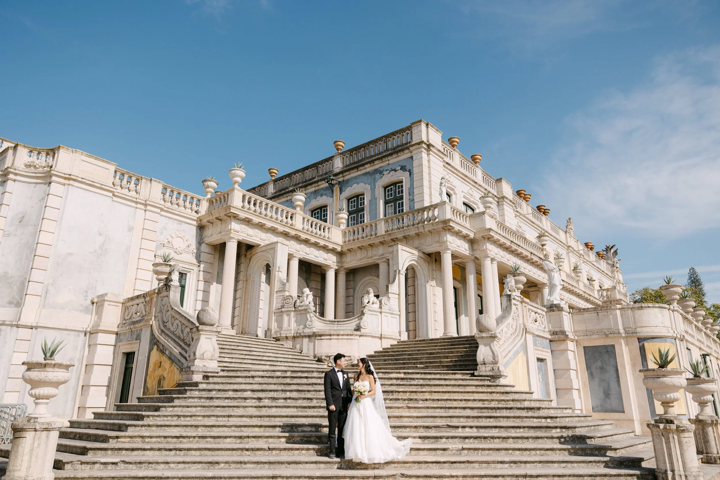 Bride and groom standing together on the grand stone staircase of Palácio Nacional de Queluz, surrounded by the palace’s historic architecture on a sunny wedding day.