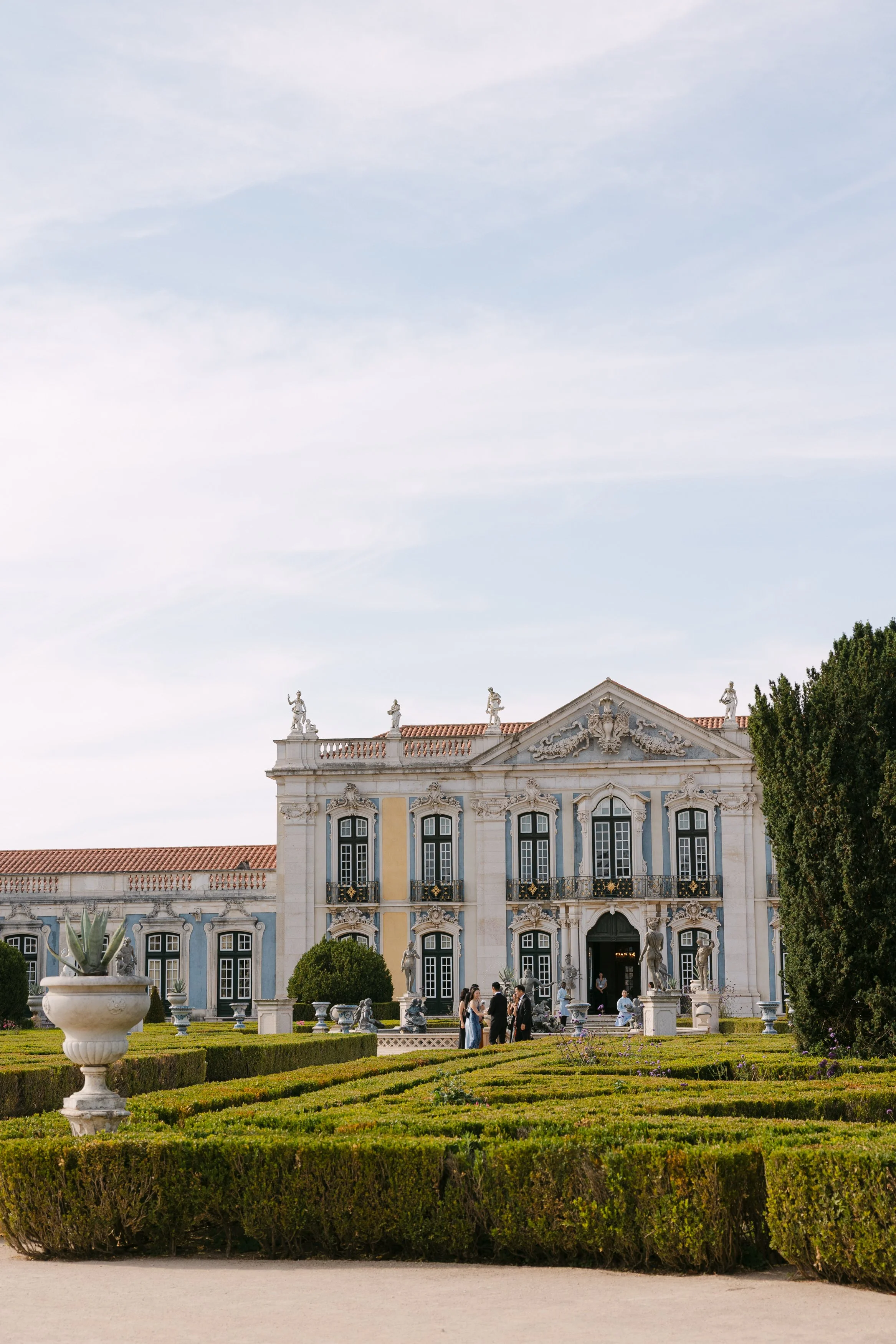 Formal gardens and historic architecture of Palácio Nacional de Queluz during a summer wedding in Portugal, with guests gathering near the palace in the distance.