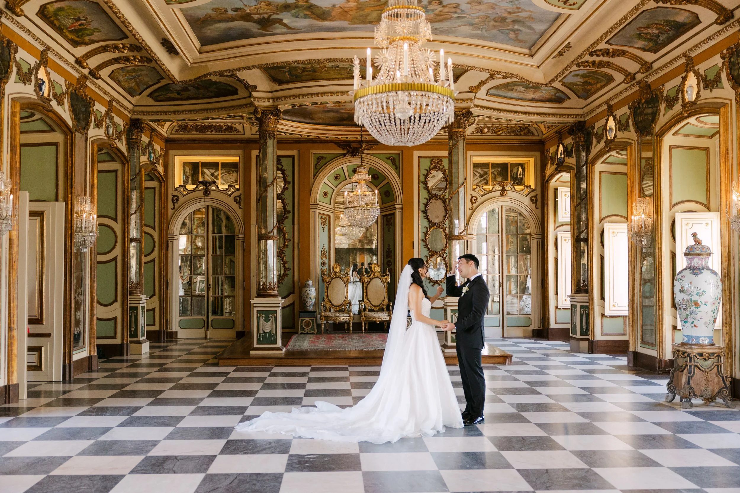 Bride and groom standing together beneath a crystal chandelier in the grand Throne Room at Palácio Nacional de Queluz, surrounded by gilded mirrors, painted ceilings and a black and white chequered floor.