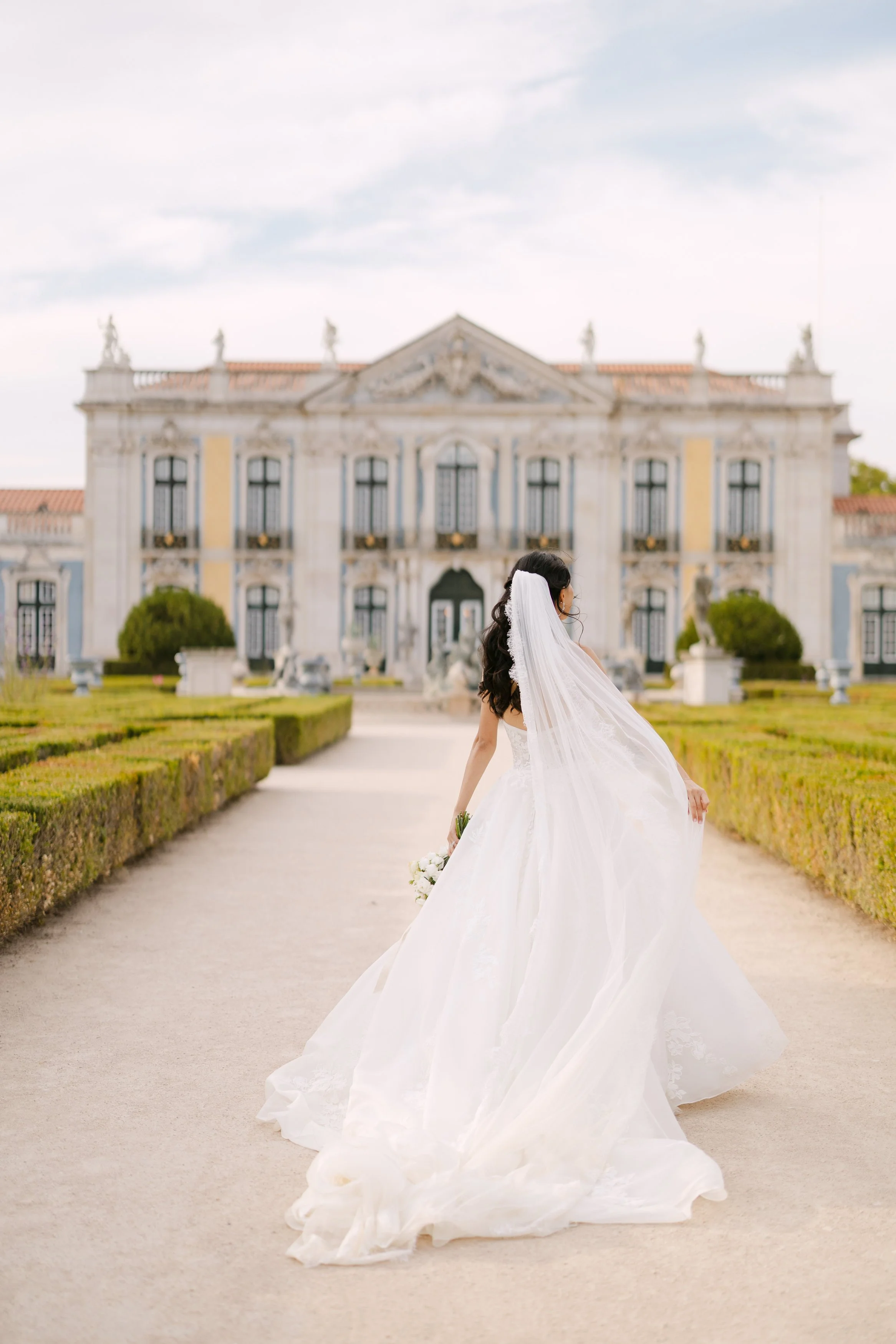 Bride walking toward Palácio Nacional de Queluz with her long wedding veil and embroidered gown flowing behind her through the palace gardens.