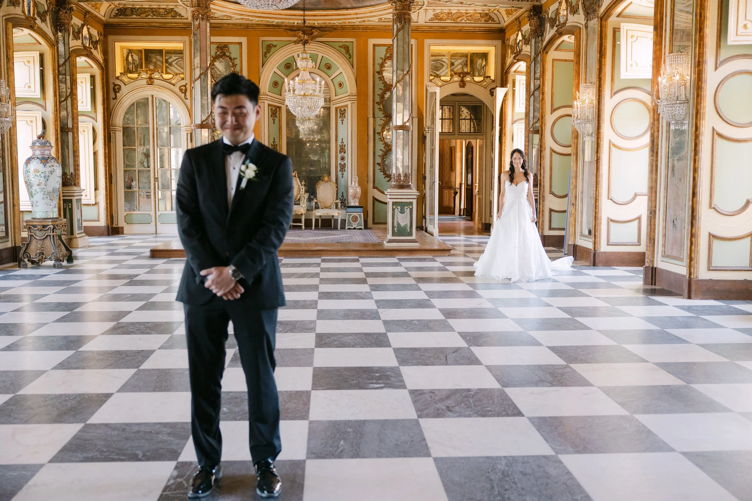 Bride approaching the groom for their first look inside an ornate palace room with a black and white chequered floor, chandeliers and historic painted details at Palácio Nacional de Queluz.