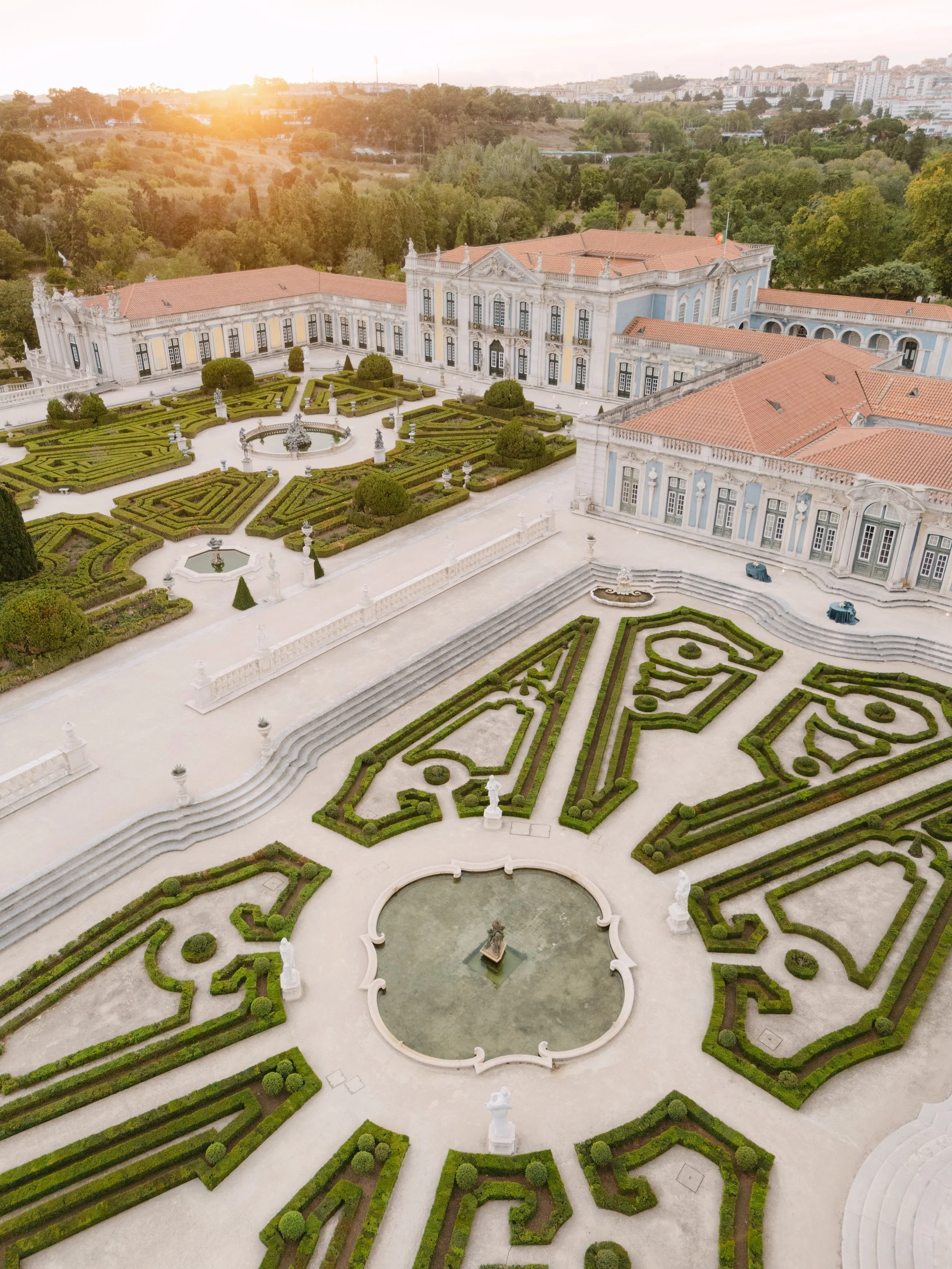 Aerial view of Palácio Nacional de Queluz and its geometric formal gardens at golden hour, showing the historic palace, fountains and surrounding landscape.