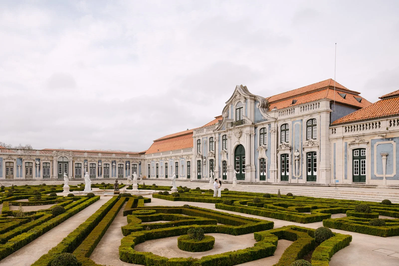 Wide view of Palácio de Queluz formal gardens with engaged couple exploring the grounds.