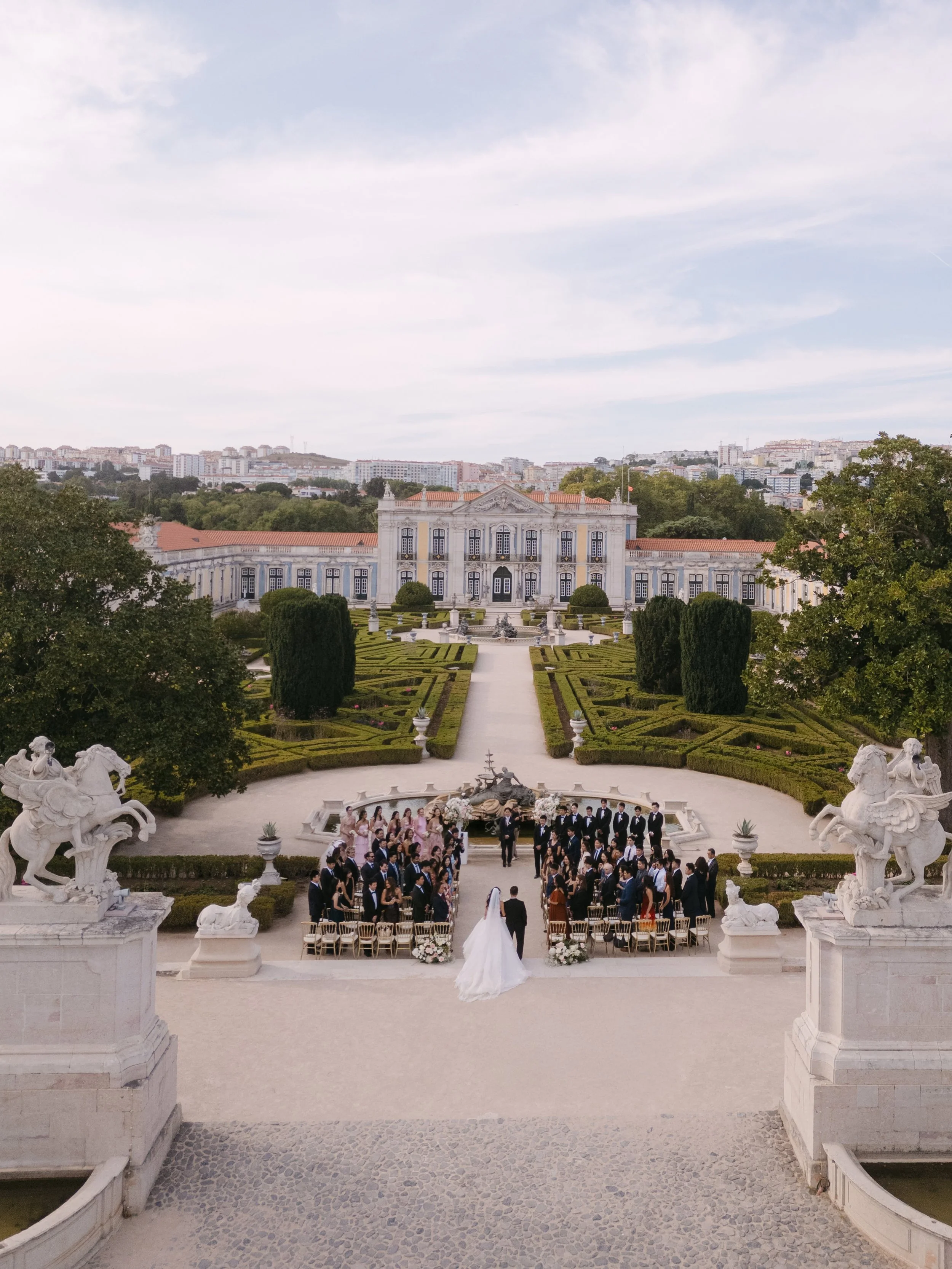 Wide view of an outdoor wedding ceremony overlooking the formal gardens and historic architecture of Palácio Nacional de Queluz in Portugal.