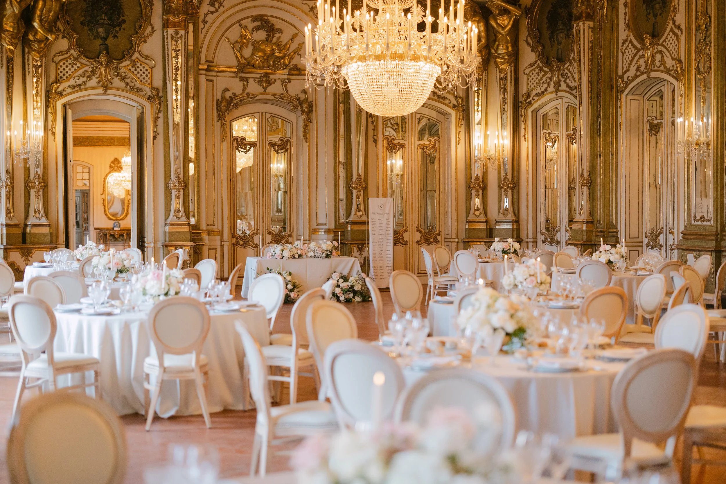 Elegant wedding reception inside an ornate ballroom at Palácio Nacional de Queluz, with round tables, soft floral arrangements, chandeliers and gilded historic interiors.