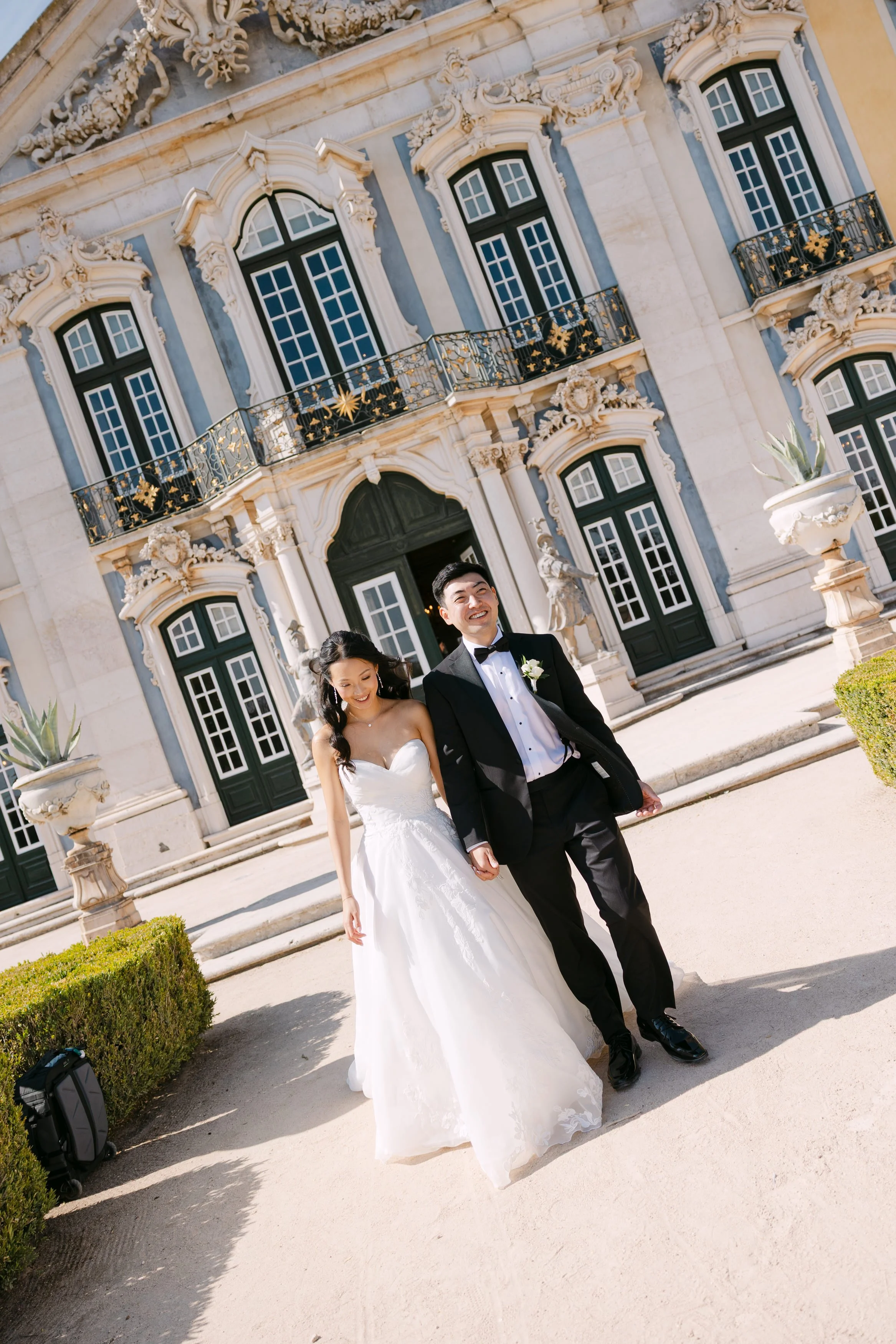 Bride and groom walking hand in hand through the formal gardens of Palácio Nacional de Queluz, with the ornate blue and white palace façade behind them.
