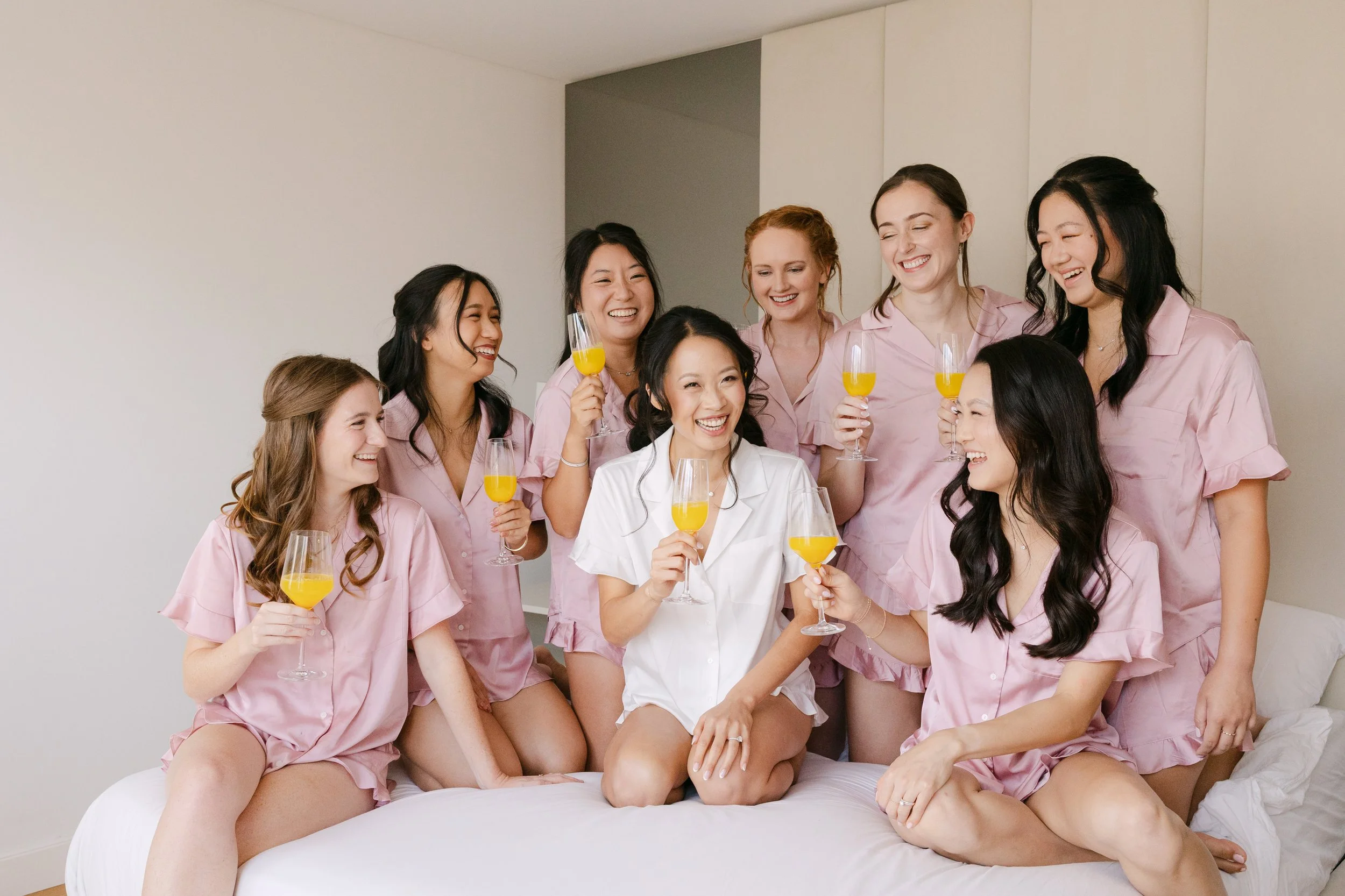 Bride in white pyjamas surrounded by her bridesmaids in matching pink pyjamas, laughing together with glasses of orange juice during wedding morning preparations.