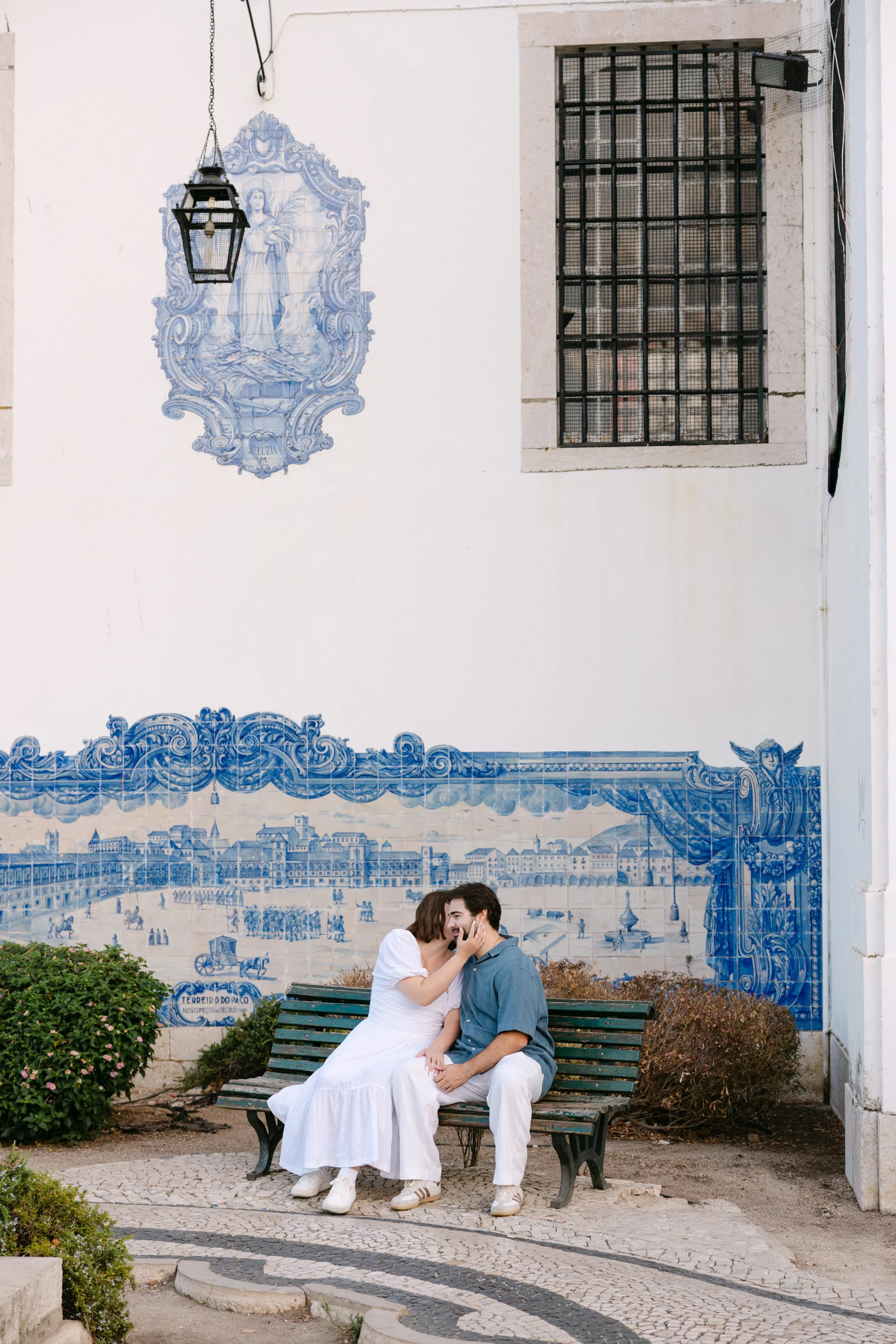 Newly engaged couple kissing on a bench beside the traditional azulejo tiles at Miradouro de Santa Luzia in Lisbon.