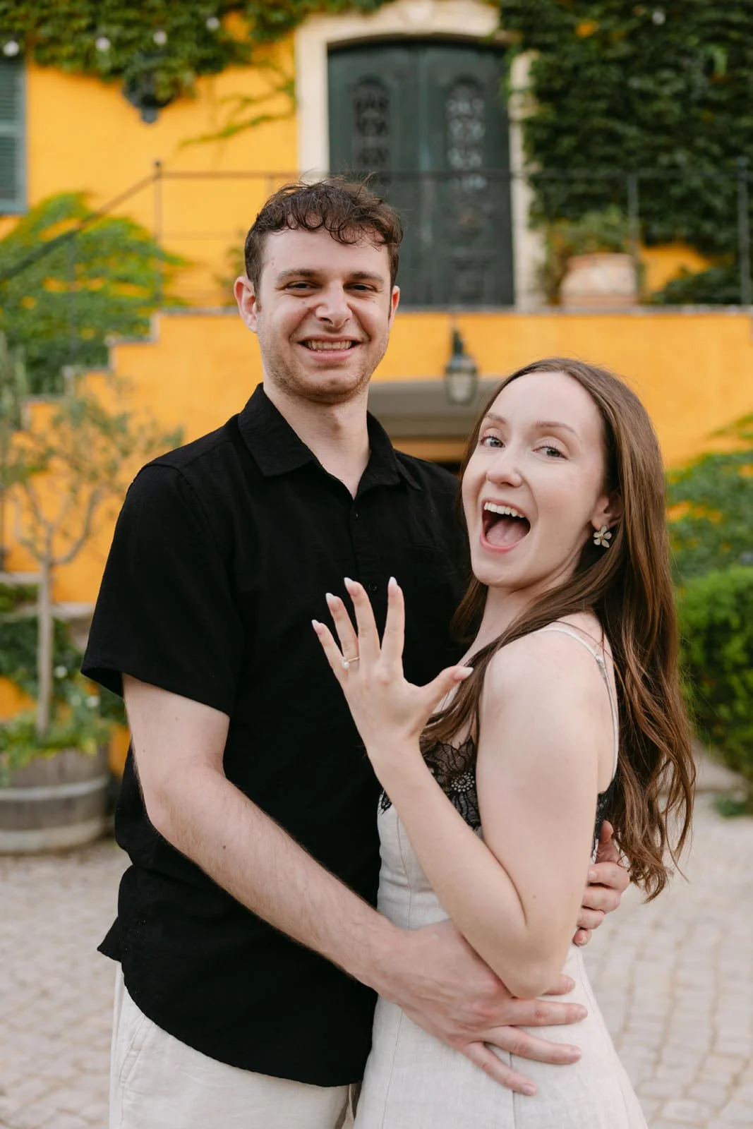 Happy newly engaged couple celebrating their surprise proposal in front of the ivy-covered Quinta de Sant’Ana manor house in Portugal.