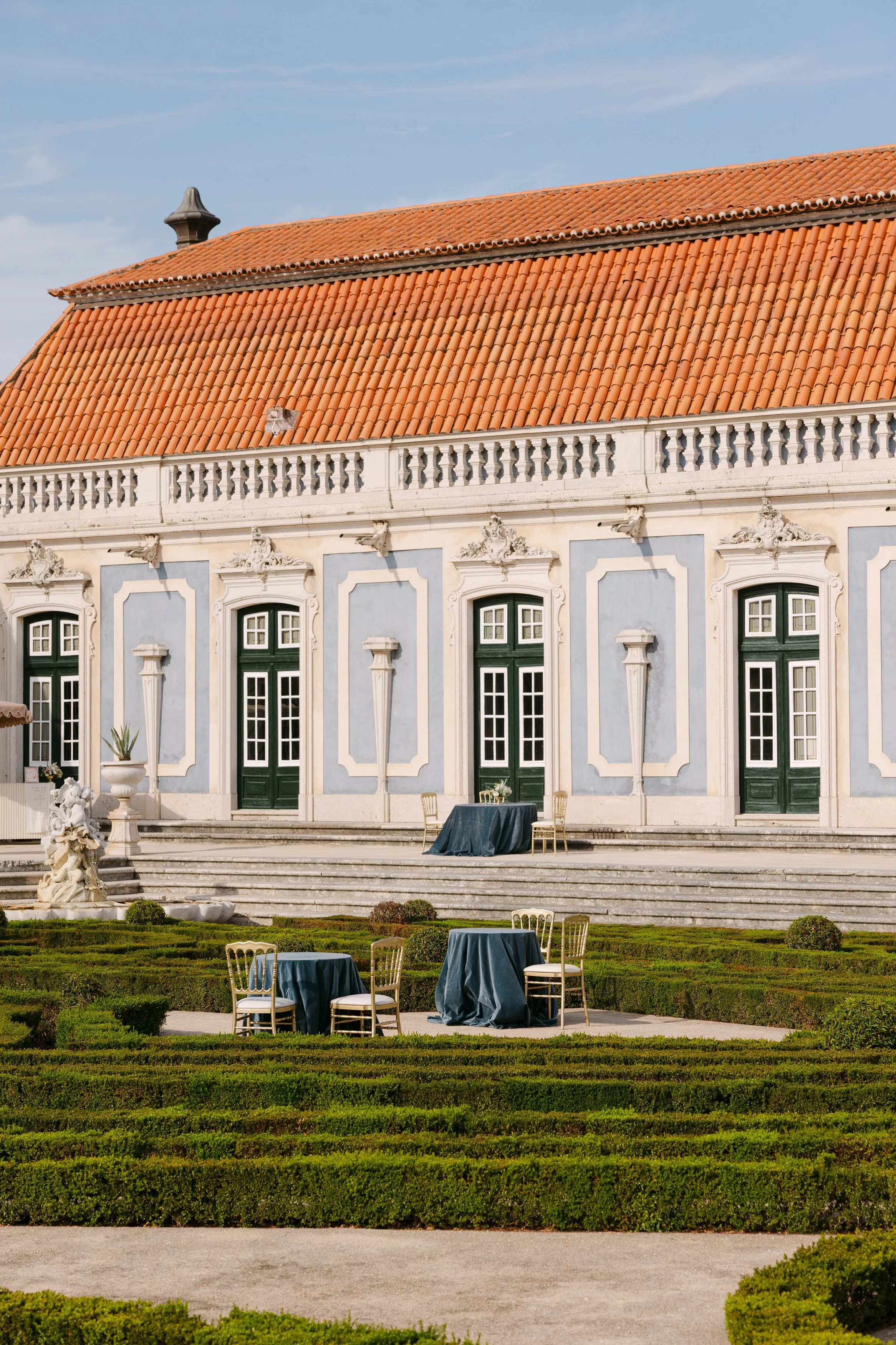 Outdoor wedding reception tables and gold chairs set among the manicured formal gardens of Palácio Nacional de Queluz, with the palace’s blue façade and terracotta roof behind them.