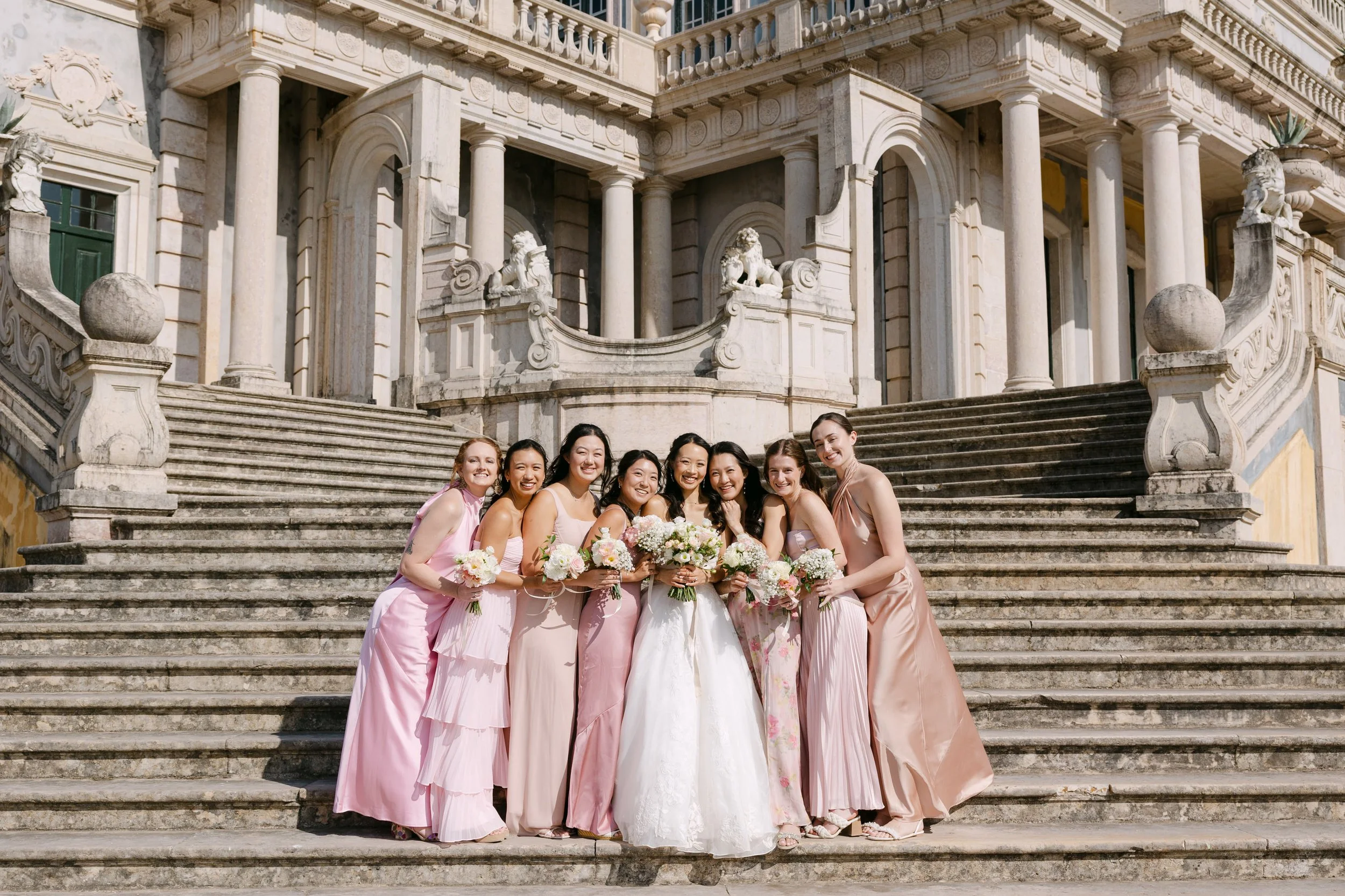 Bride surrounded by her bridesmaids in different shades of pink, holding pastel bouquets and smiling together on the grand staircase of Palácio Nacional de Queluz.