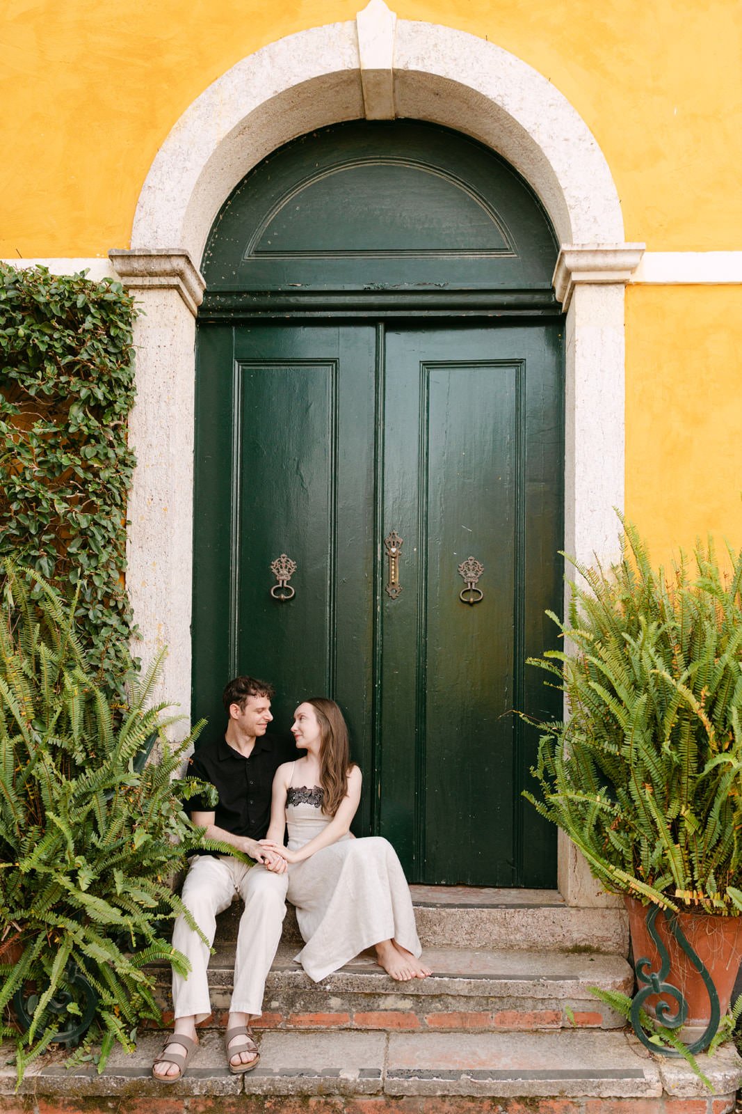 Engaged couple sitting hand in hand beneath a large green doorway surrounded by ferns at Quinta de Sant’Ana in Portugal.