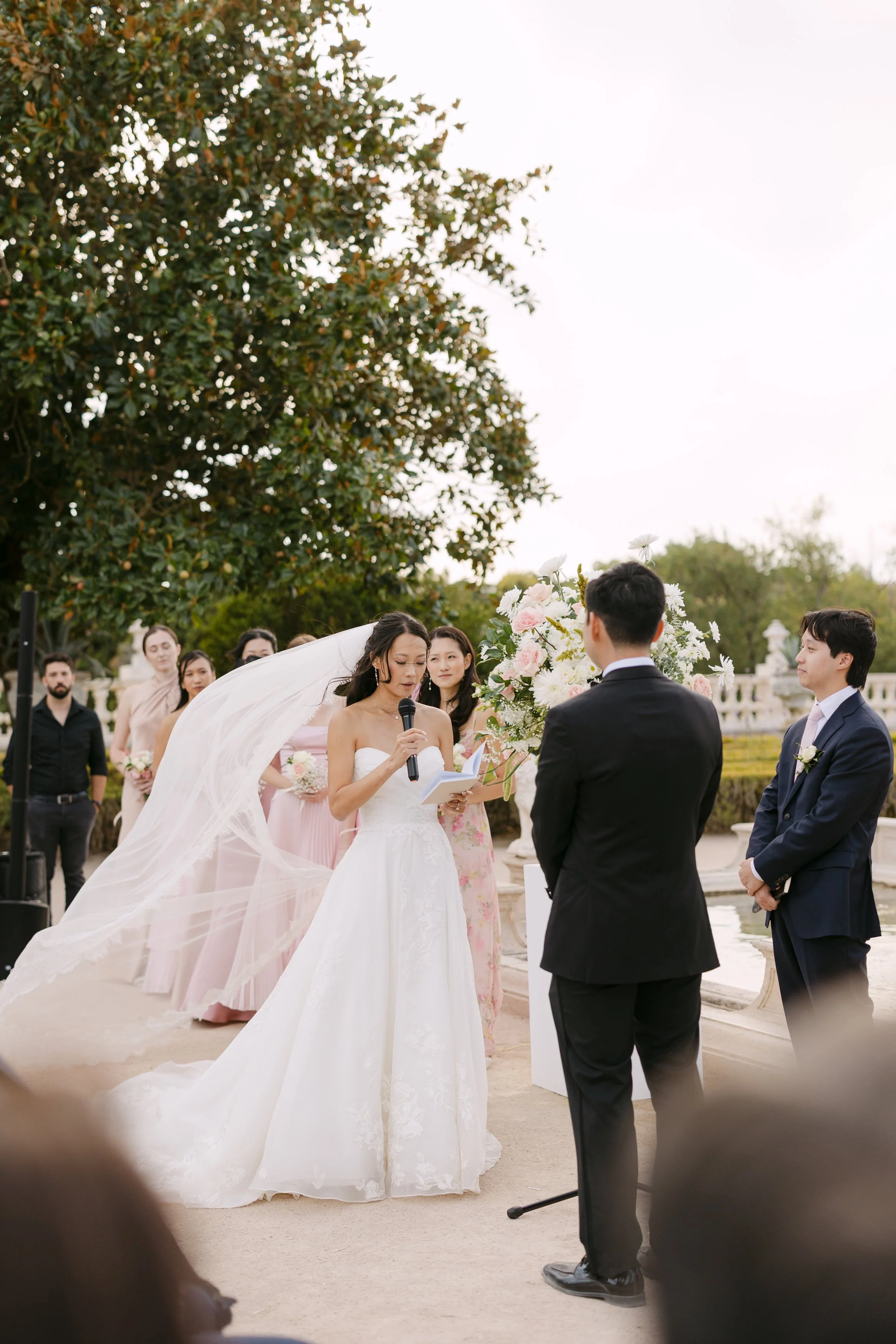 Bride reading her vows to the groom during their outdoor wedding ceremony, with her veil blowing in the breeze and pastel flowers behind them at Palácio Nacional de Queluz.