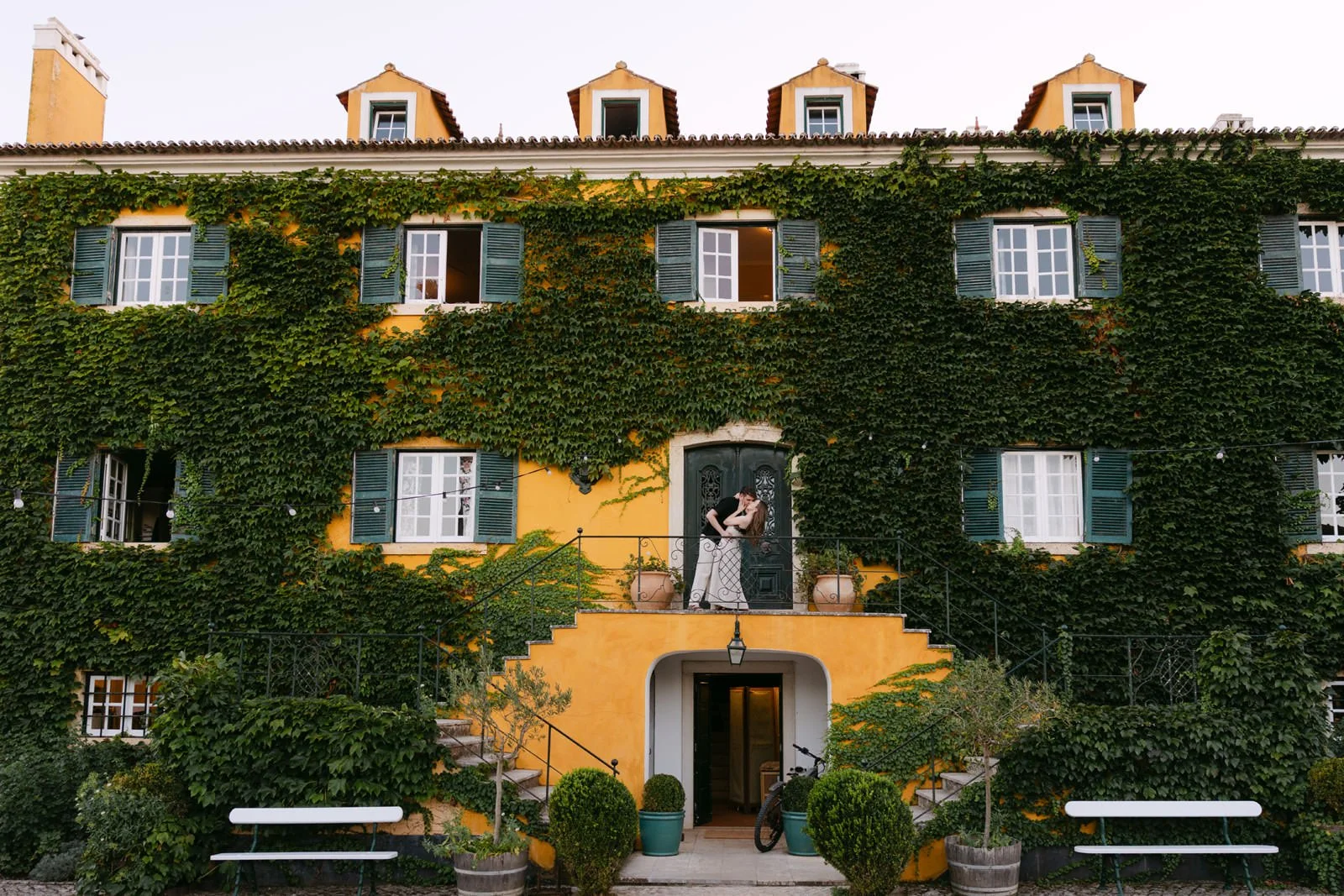 Engaged couple embracing on the steps of the ivy-covered yellow manor house at Quinta de Sant’Ana in Portugal.