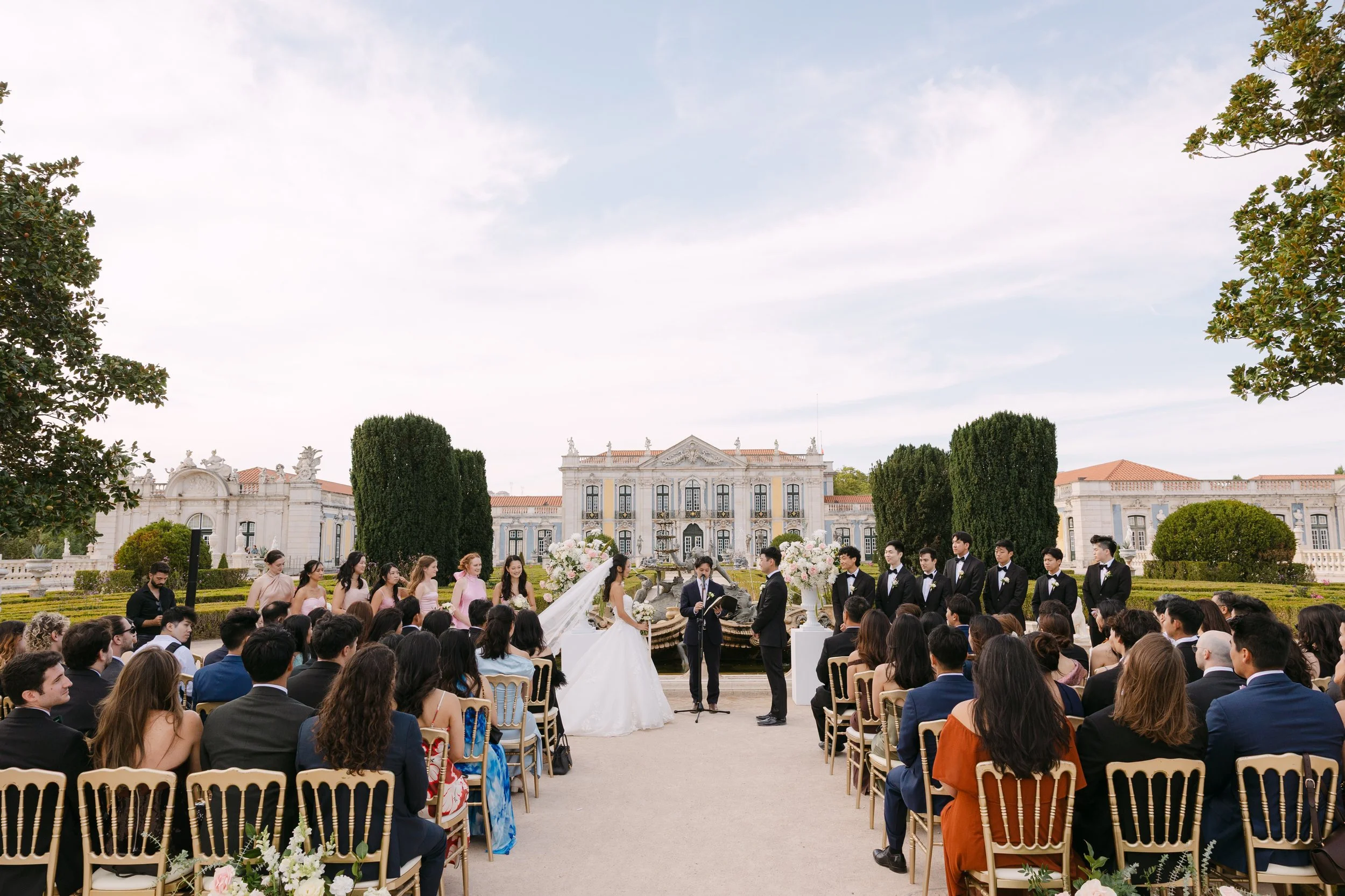 Bride and groom standing together during their outdoor wedding ceremony, surrounded by bridesmaids, groomsmen, pastel flowers and guests with Palácio Nacional de Queluz in the background.