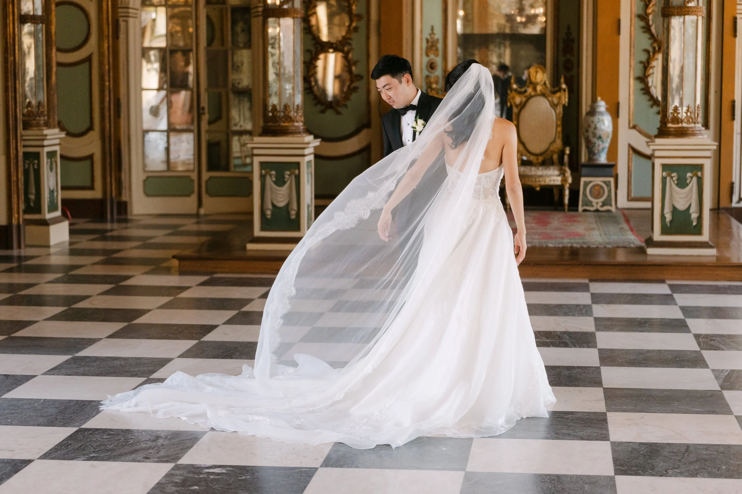 Bride in a flowing white wedding gown with her long veil sweeping through the air as the groom stands behind her inside Palácio Nacional de Queluz.