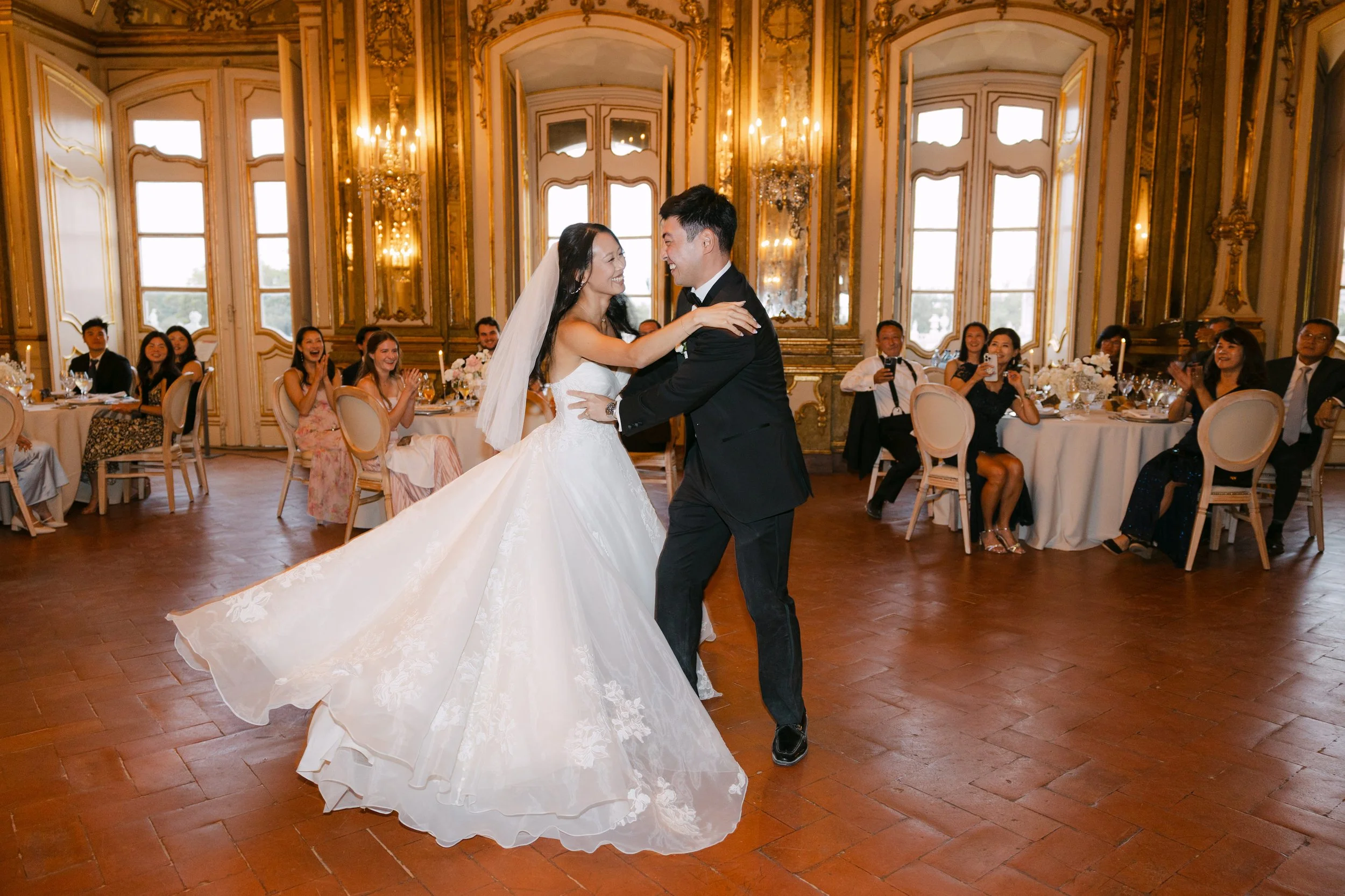 Bride and groom laughing during their first dance inside the ornate ballroom at Palácio Nacional de Queluz, surrounded by their wedding guests.