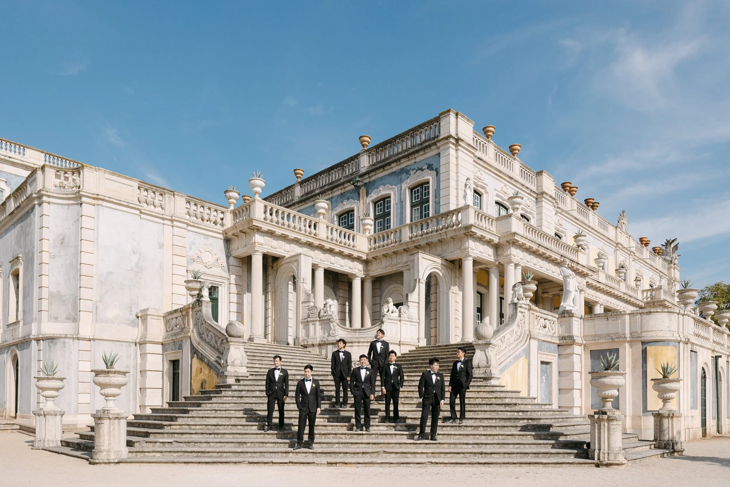 Groom and groomsmen in black tuxedos posing across the grand stone staircase outside Palácio Nacional de Queluz on a sunny wedding day.