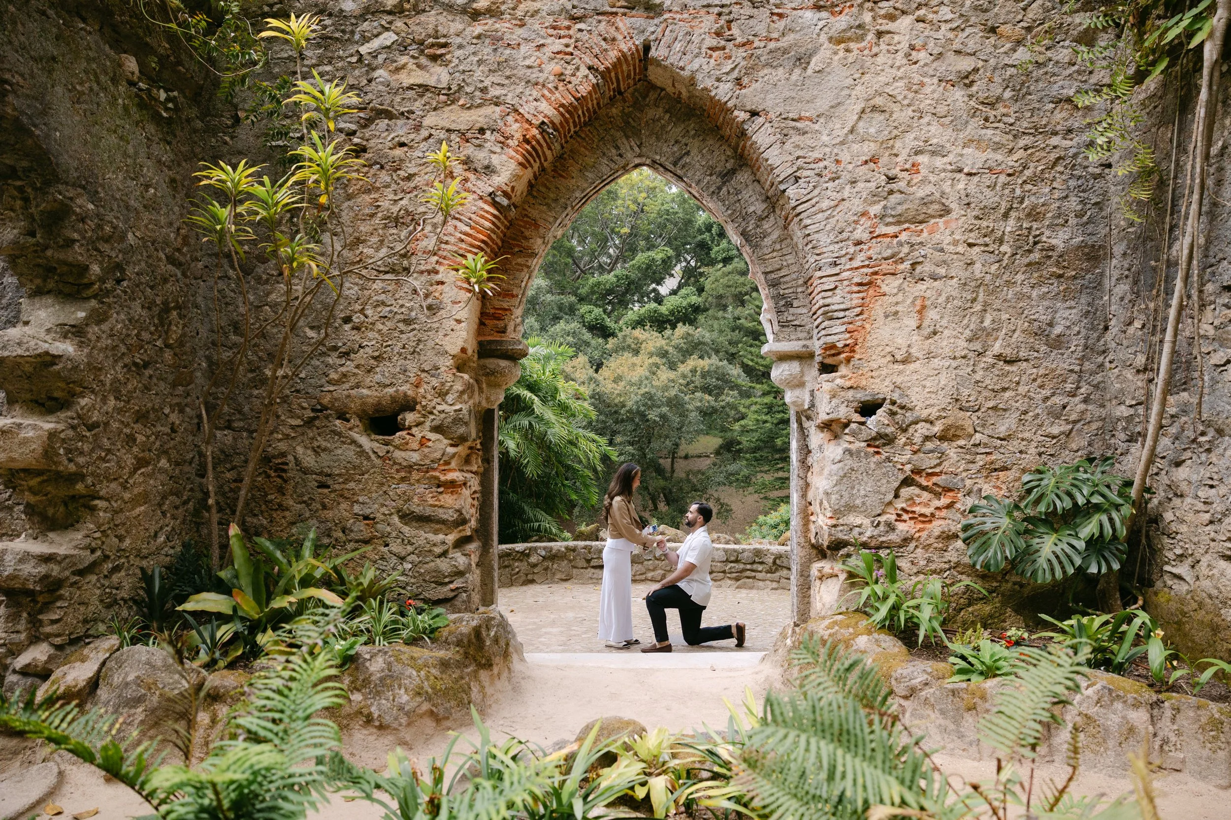 Couple during a surprise proposal at the Monserrate ruins in Sintra surrounded by historic stone arches and greenery.