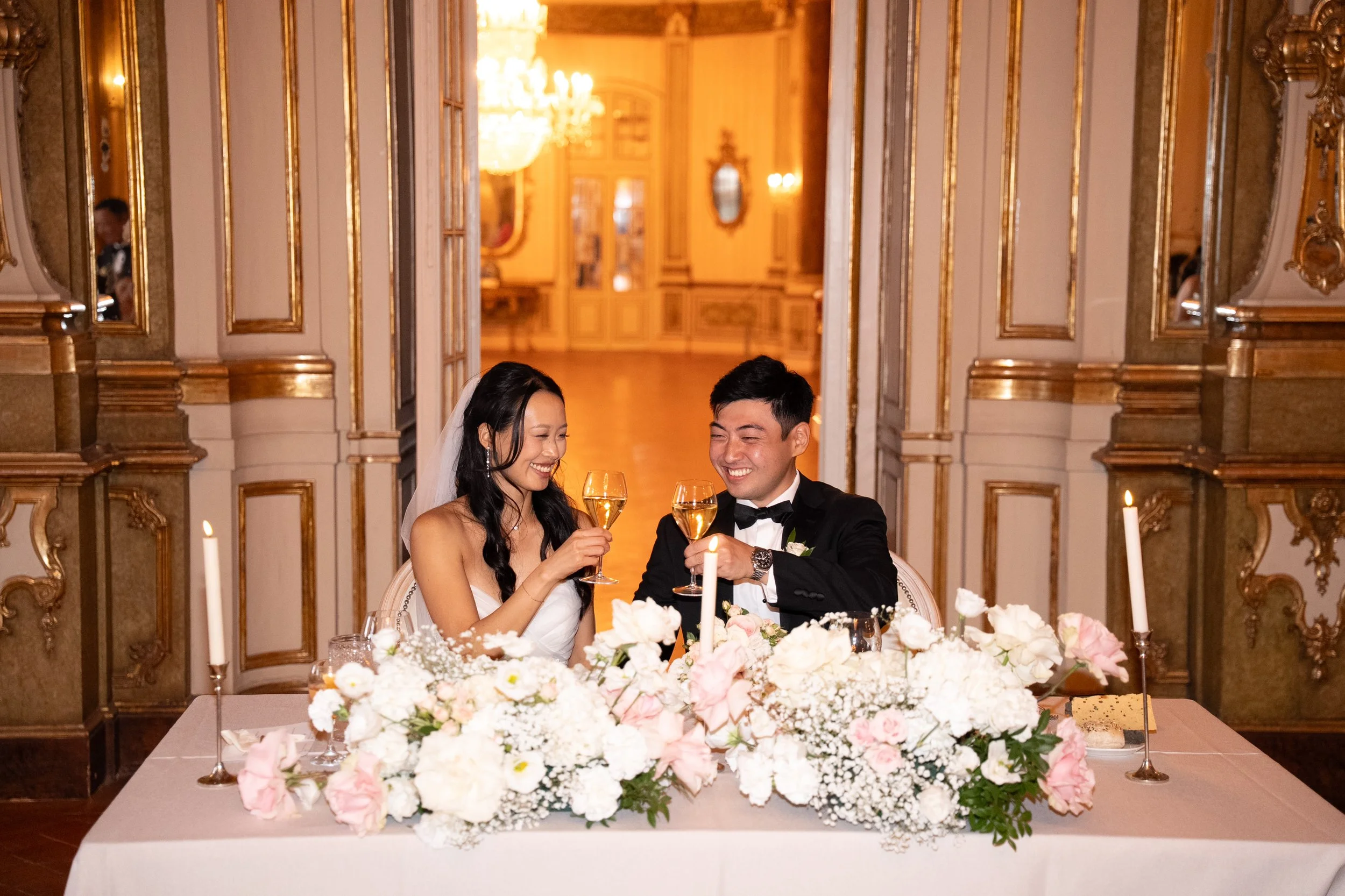Newlyweds smiling and raising their glasses at a flower-filled sweetheart table during their wedding reception at Palácio Nacional de Queluz.
