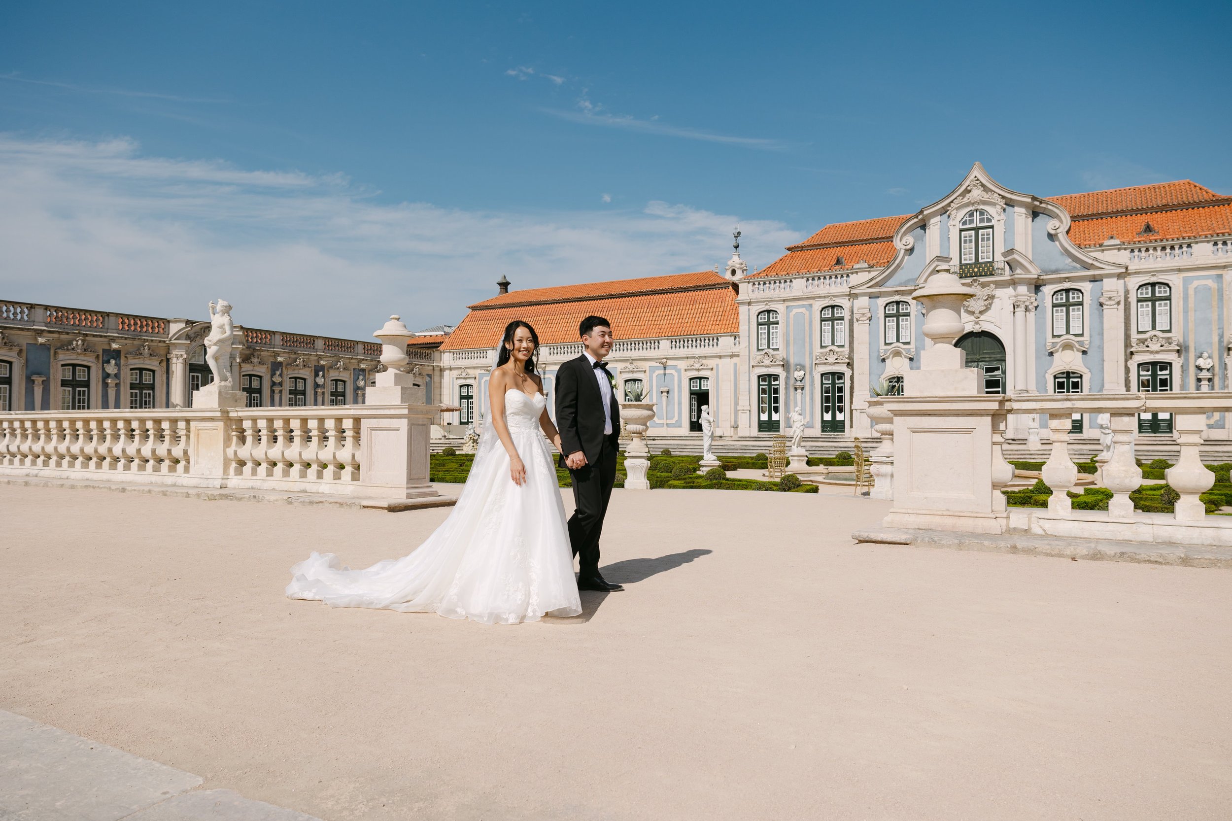 Bride and groom holding hands in front of Palácio Nacional de Queluz on a sunny wedding day, surrounded by the palace’s historic architecture and formal gardens.