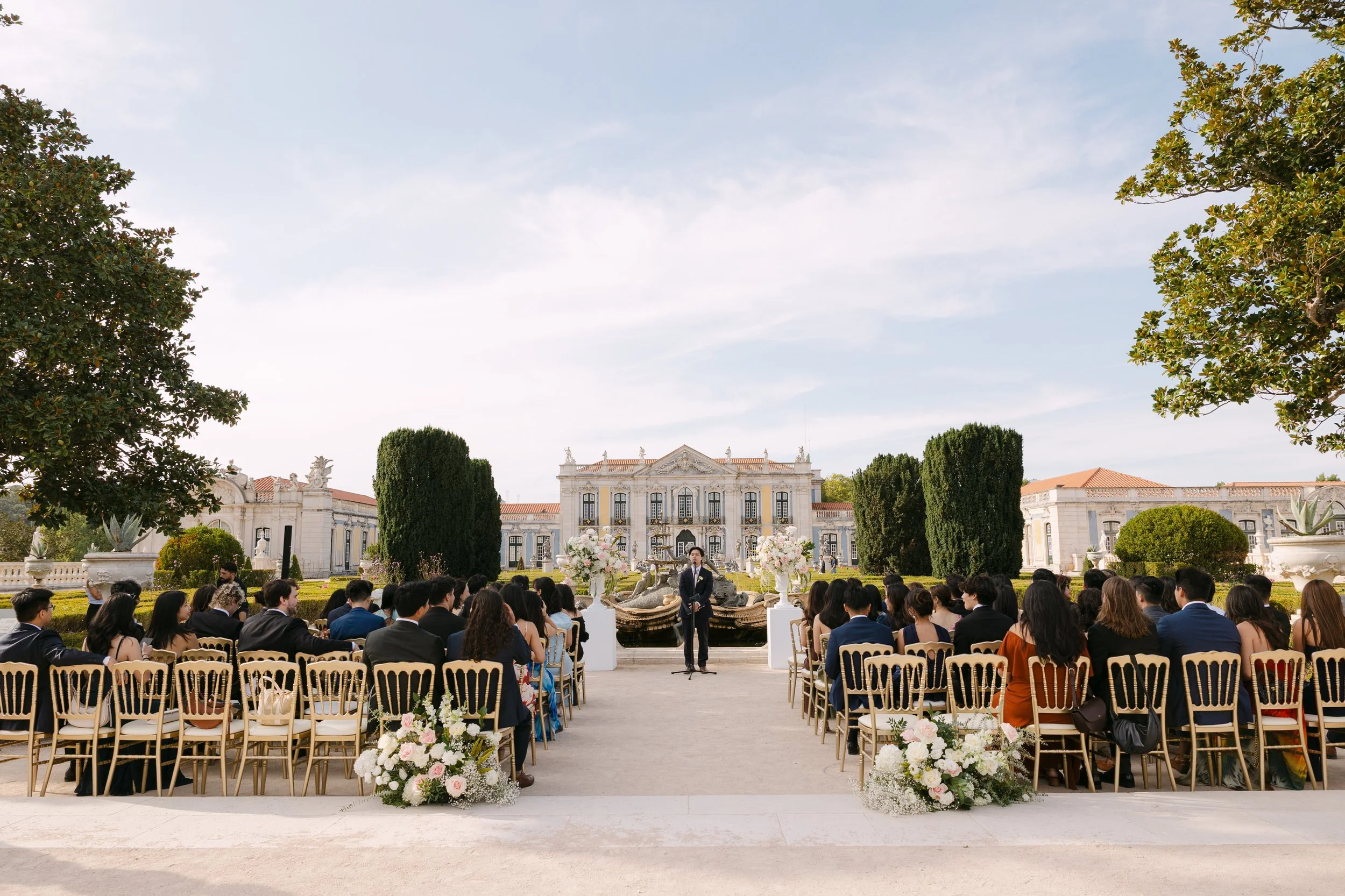 Outdoor wedding ceremony in the formal gardens of Palácio Nacional de Queluz, with guests seated on gold chairs facing the floral altar and historic palace in the background.