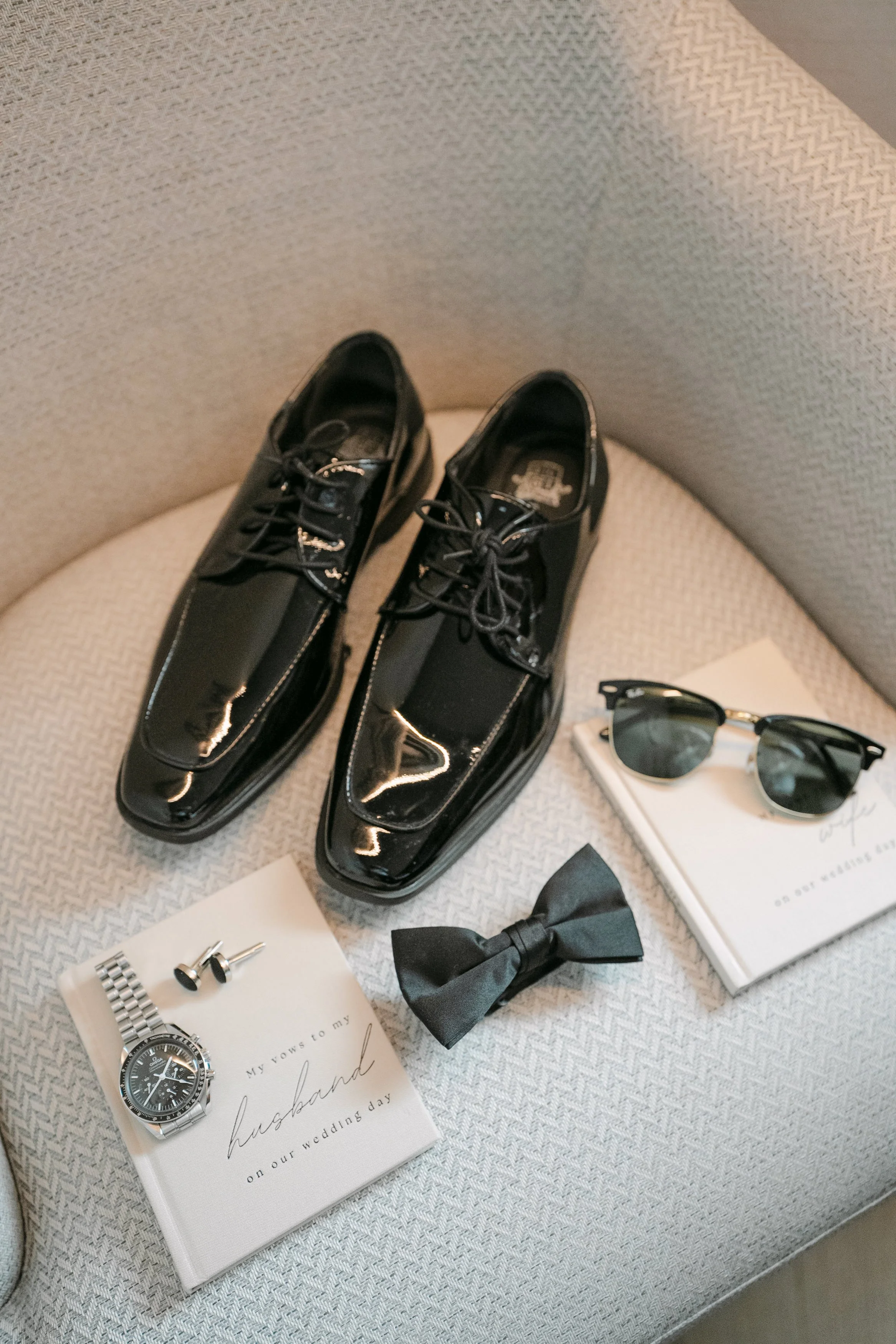 Black patent groom shoes styled with a black bow tie, silver watch, cufflinks, sunglasses and a personalised wedding-day vow book during morning preparations for a Lisbon wedding.