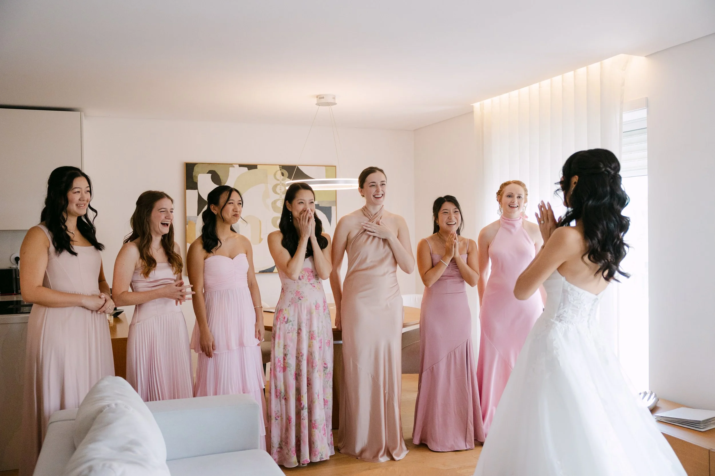 Bridesmaids in different shades of pink reacting with smiles and happy surprise as they see the bride in her wedding dress for the first time.