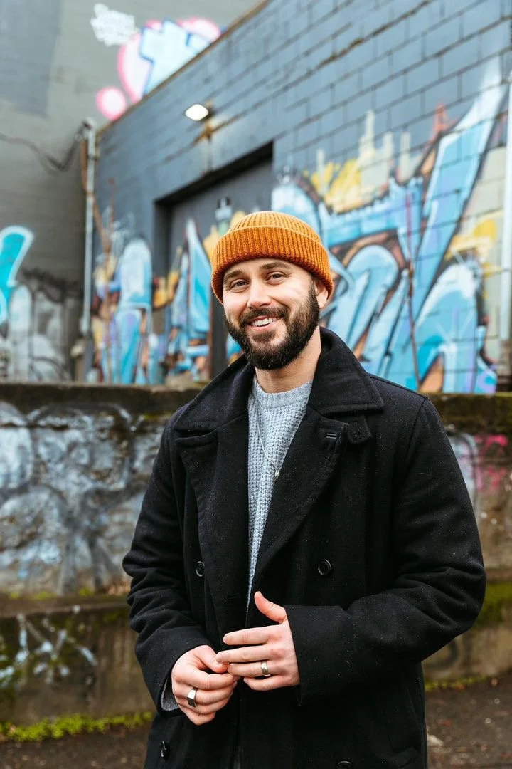 James, a trans man who is a motivational speaker and stress management coach, who coaches you through your stressful life transition, smiles while in a beanie and in front of a graffiti wall.