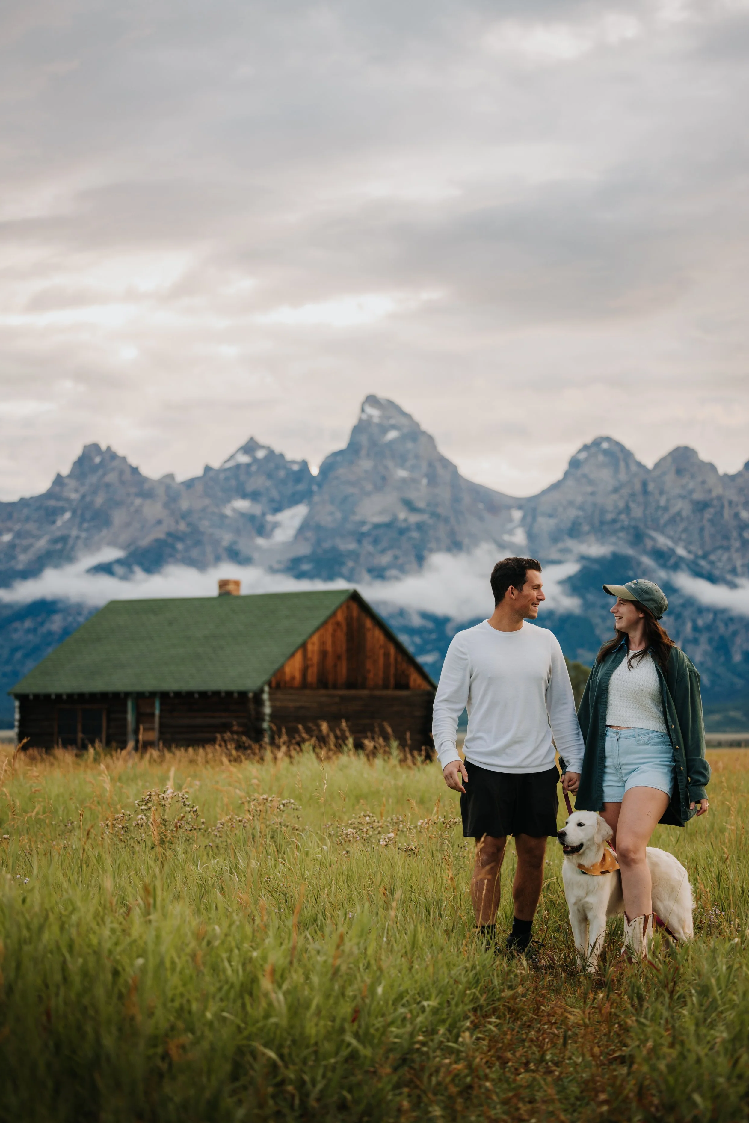 A couple walking with their dog in a grassy field with mountains in the background and a wooden cabin of green roof.
