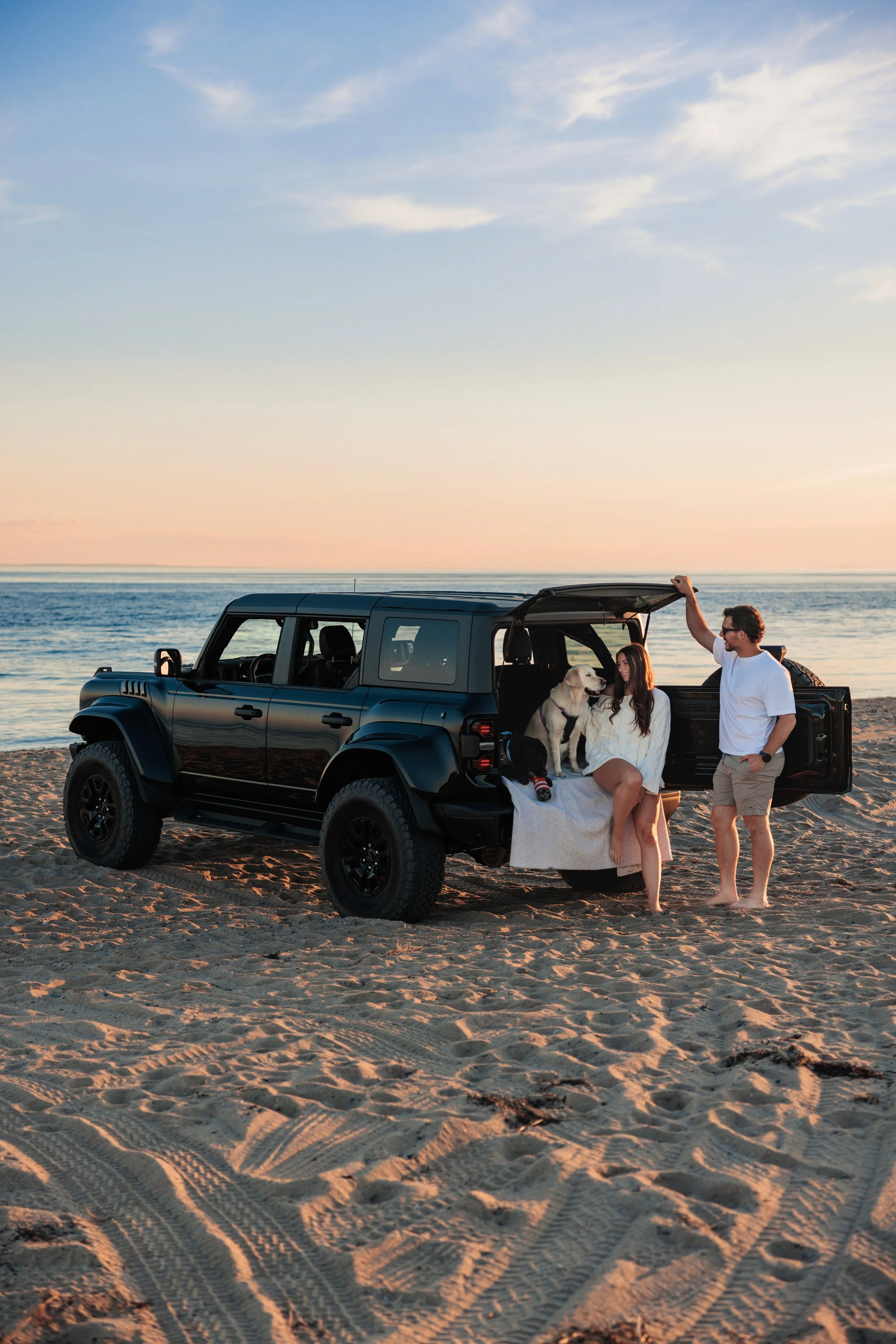 A family with a dog stands near a black SUV on a beach at sunset, with the ocean and sky in the background.