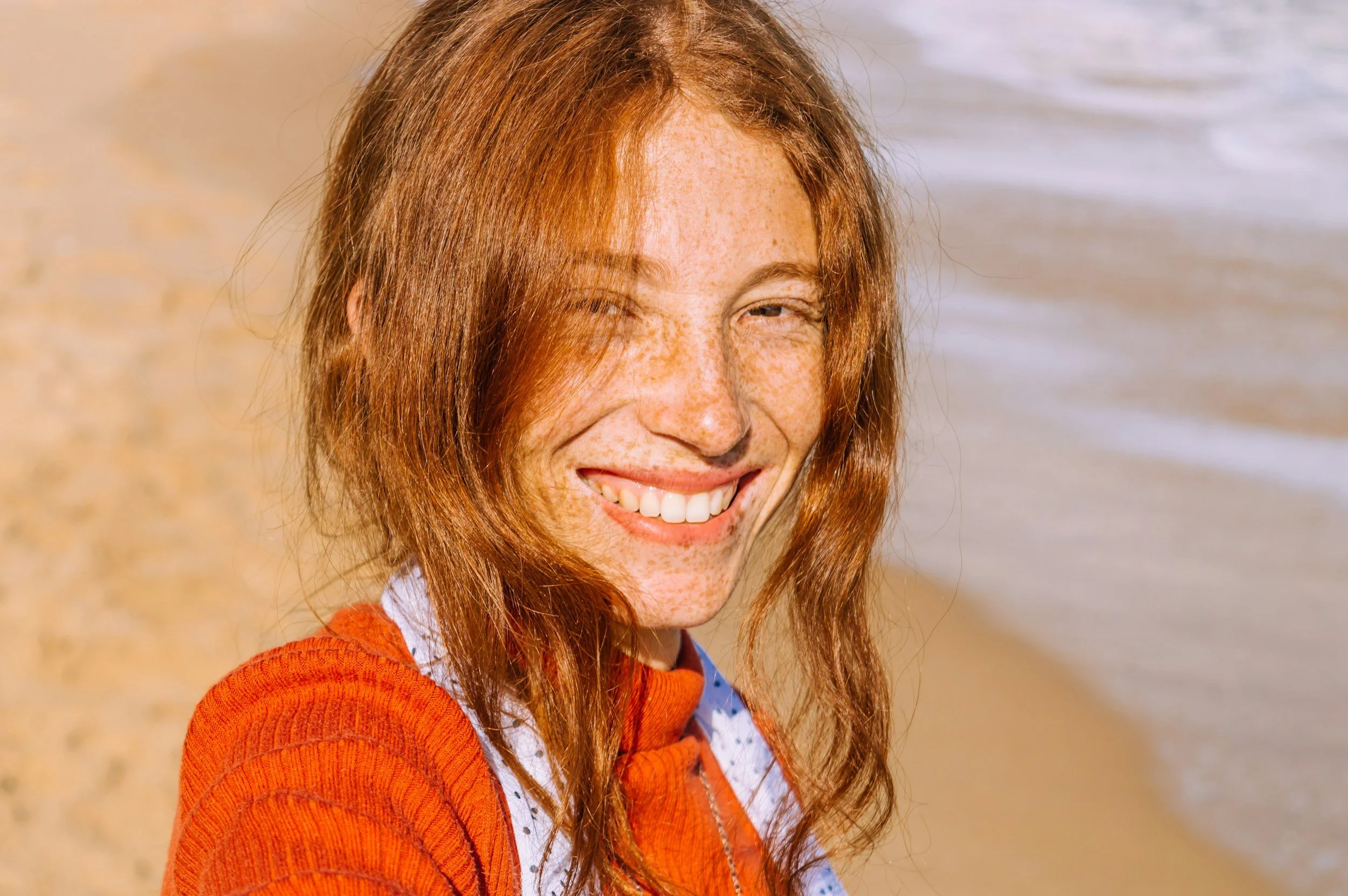 woman with red hair and freckles smiling in the sun with the beach and ocean in the background